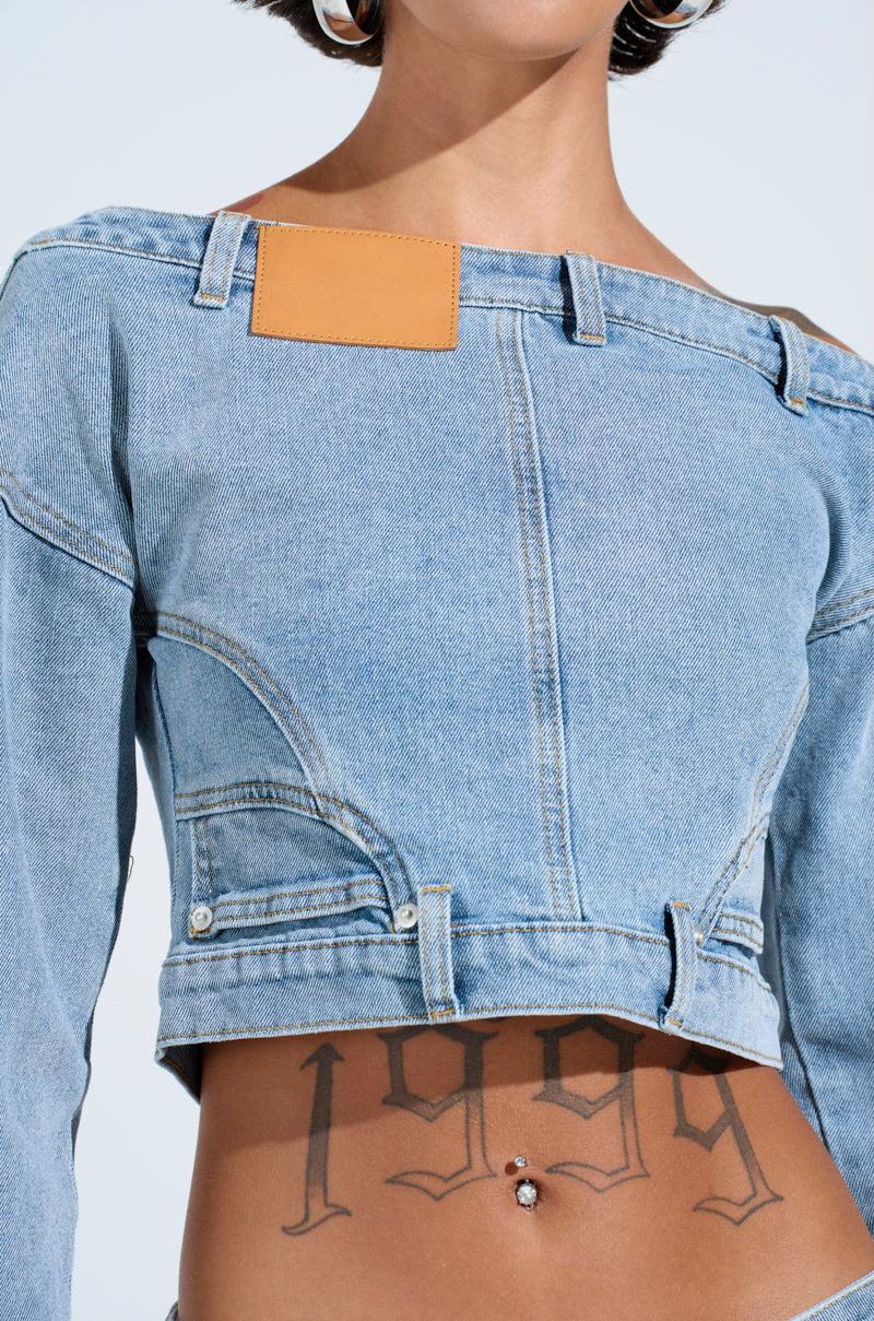 Wearing the SHAKE IT OFF DENIM OFF THE SHOULDER TOP with tan patches, a person reveals a "1999" tattoo with an arrow on their lower abdomen. The look is finished with a belly button piercing and hoop earrings, showcasing effortless style.