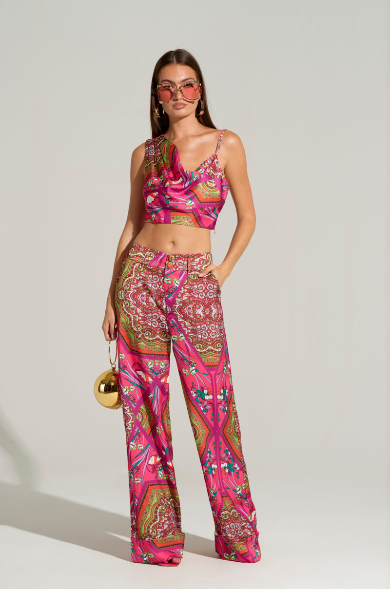 A woman stands confidently wearing a vibrant AKIRA Label MOVE AND SHAKE TROUSER IN PINK MULTI sleeveless top and matching wide-leg pants. She accessorizes with pink-tinted sunglasses and holds a small round gold handbag. The plain background highlights her stylish outfit.