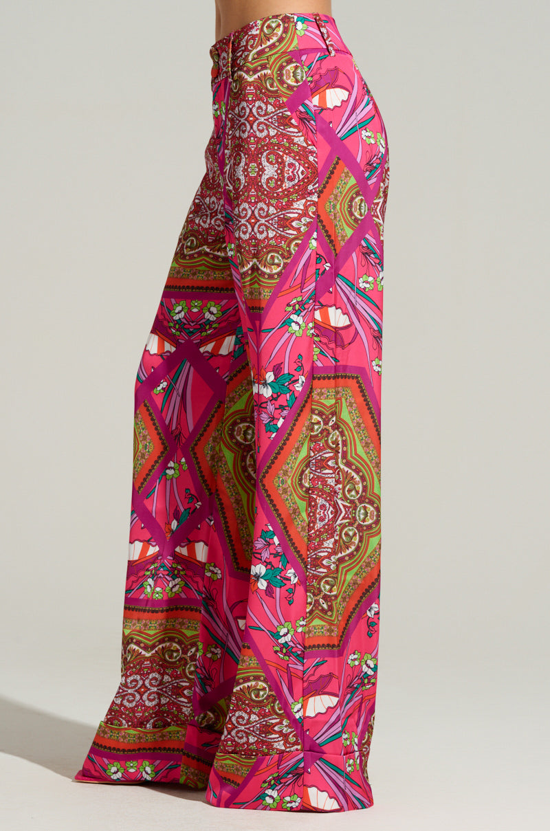 A person is wearing the MOVE AND SHAKE TROUSER IN PINK MULTI from AKIRA Label, featuring wide-leg pants adorned with a bold geometric and floral print in vibrant shades of pink, green, and white. The colorful pattern contrasts strikingly against a neutral background, capturing a retro style that is perfect for making a statement.