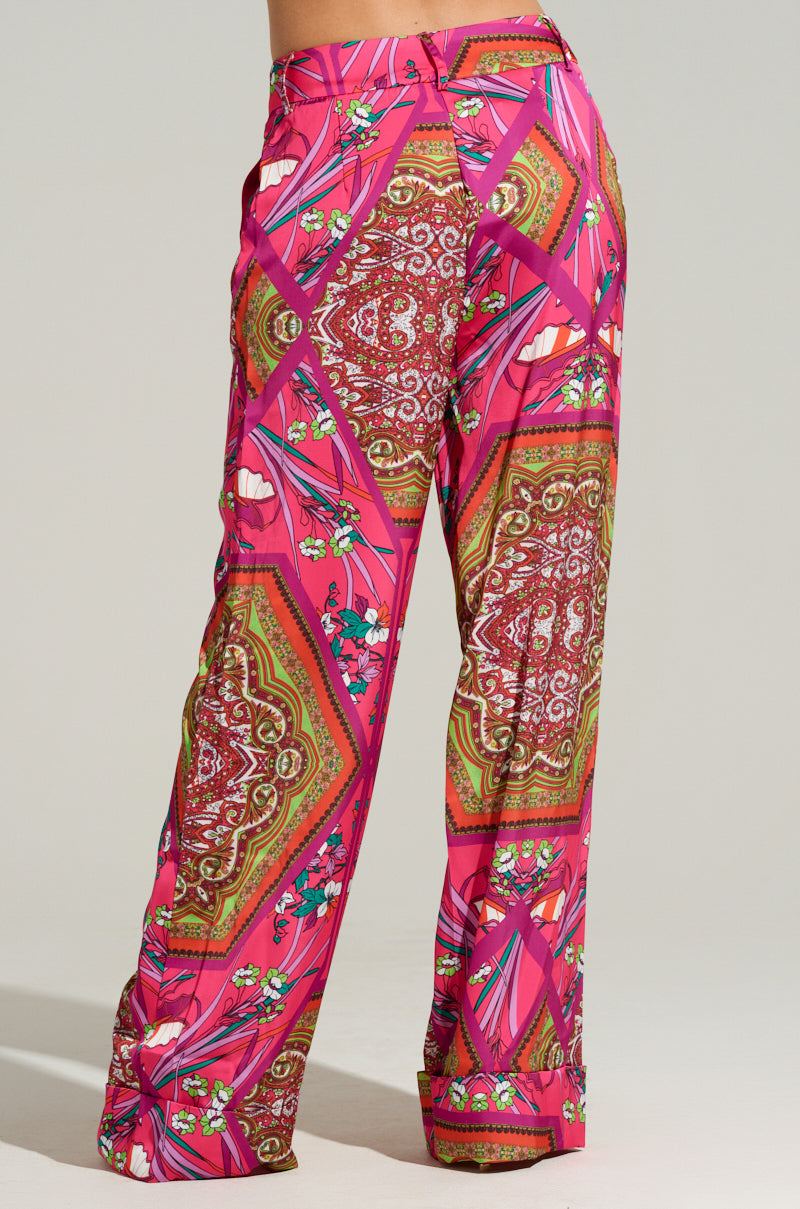 A person wearing the MOVE AND SHAKE TROUSER IN PINK MULTI from the AKIRA Label showcases brightly patterned pink and multicolored palazzo pants with intricate paisley designs and floral motifs. The wide-leg silhouette and high waist design contribute to a vibrant, stylish look.