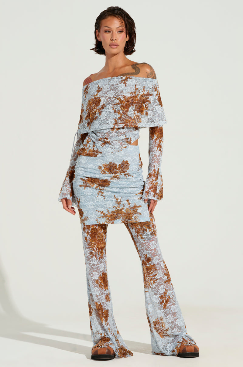 A person dons a light blue and brown floral off-the-shoulder top paired with the GIVING YOU SOMETHING TO DREAM ABOUT PANT, which features stretchy lace knit material and a skirt overlay. They stand confidently against a plain background, showcasing their stylish outfit.