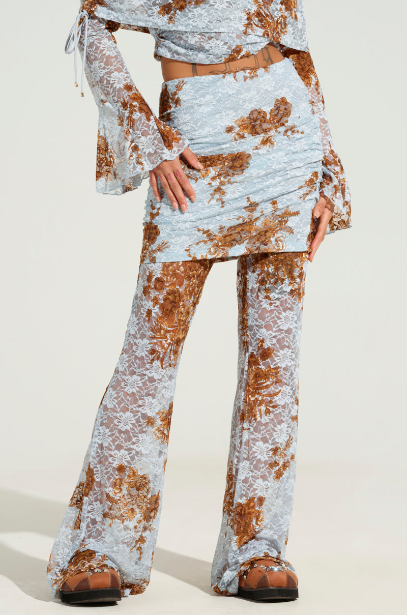 Dressed in a chic two-piece outfit featuring floral lace patterns in brown and blue, the ensemble includes a flared top, a mini skirt, and semi-sheer pants known as the "Giving You Something to Dream About Pant." The look is finished with stylish brown platform sandals.