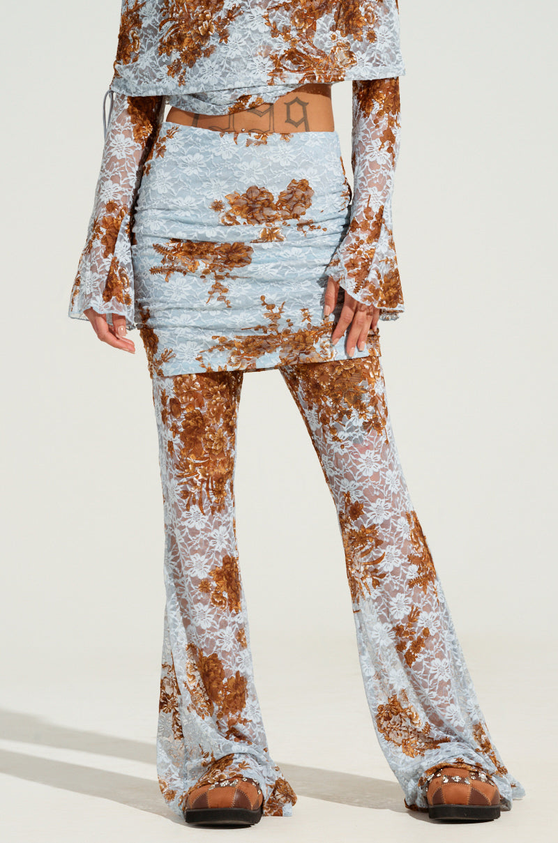 A person models the GIVING YOU SOMETHING TO DREAM ABOUT PANT, a floral-patterned two-piece ensemble featuring long sleeves and semi-sheer, flared pants in an elegant blend of light blue and brown. Brown sandals and a coordinating belt accentuate the outfit. The photo emphasizes the detailed craftsmanship of the ensemble.