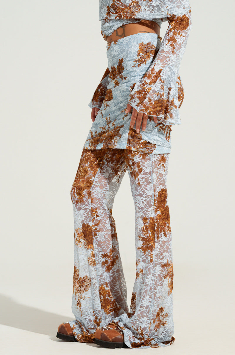 A person wearing the "Giving You Something to Dream About Pant," featuring a floral-patterned outfit with a long-sleeved top and matching semi-sheer pants, paired with heeled sandals. The ensemble showcases a light blue base adorned with brown floral designs against a plain white background.