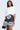 A woman wears the AKIRA Label 8BALL RHINESTONE OVERSIZED TEE, a white oversized t-shirt dress with a bold black 8-ball graphic, ruffled polka dot and striped hem, and holds a small black handbag.