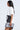 A woman with long dark hair stands sideways, wearing an AKIRA Label 8BALL RHINESTONE OVERSIZED TEE as a dress with a black 8-ball design, ruffled hem with polka dots and stripes, and holds a matching black and white clutch.