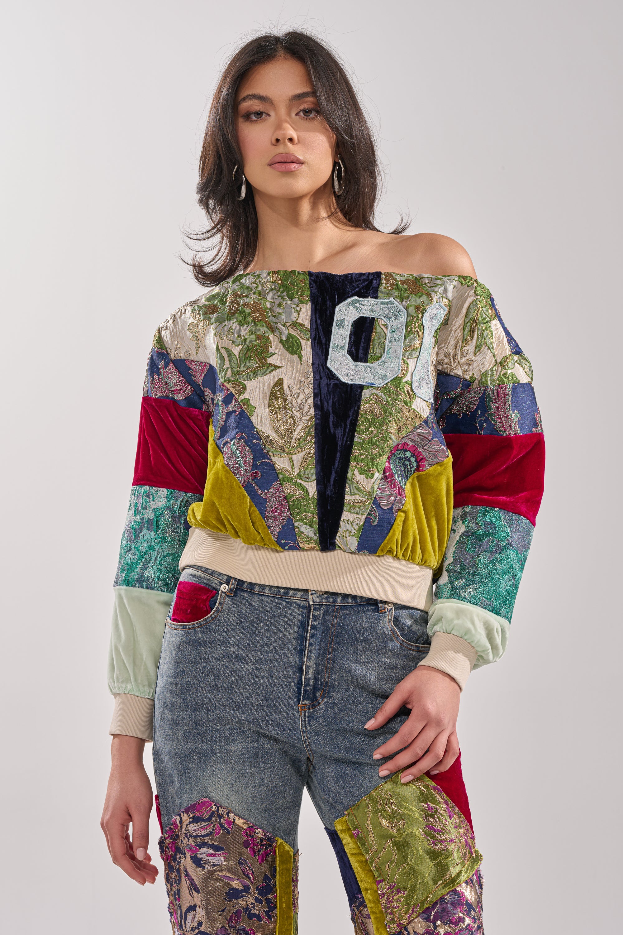 A woman wears the MIXED EMOTION BROCADE PATCHWORK SWEATSHIRT—an off-shoulder, cropped, colorful patchwork style with long sleeves—paired with blue jeans with floral fabric patches, standing against a plain light background.