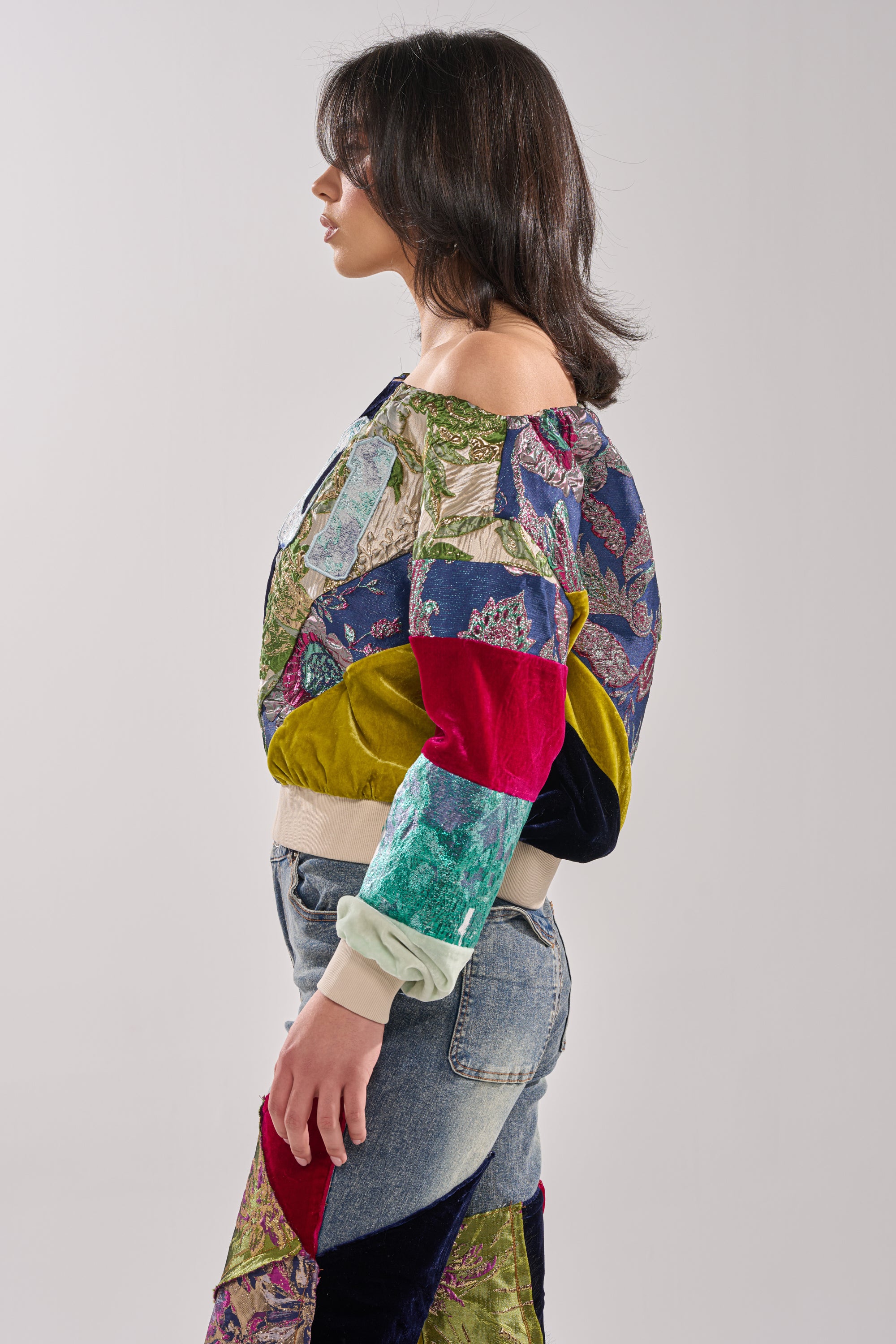 A woman in profile wears the MIXED EMOTION BROCADE PATCHWORK SWEATSHIRT with vibrant patterns, paired with light blue patchwork denim jeans. The backdrop is a simple, light gray.