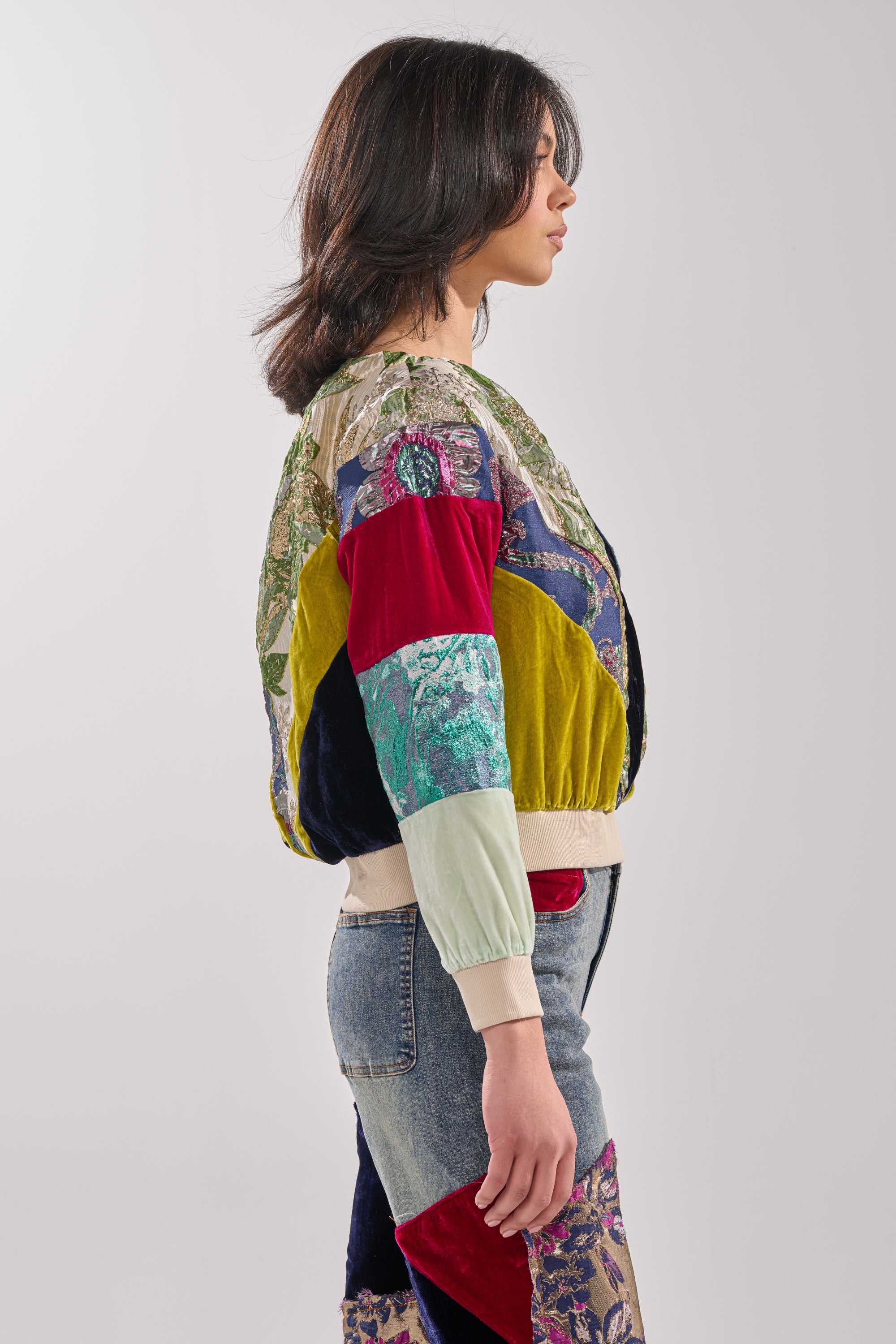 A person with shoulder-length dark hair stands in profile wearing the MIXED EMOTION BROCADE PATCHWORK SWEATSHIRT, featuring colorful patterns and textures, against a plain light background.
