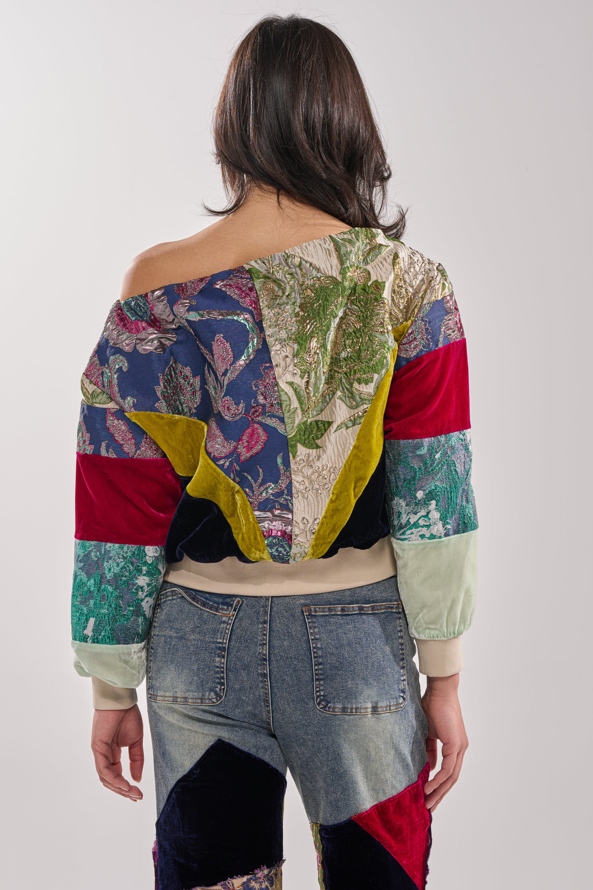 A person with shoulder-length dark hair faces away, wearing the MIXED EMOTION BROCADE PATCHWORK SWEATSHIRT off one shoulder and blue jeans with mixed fabric patches.