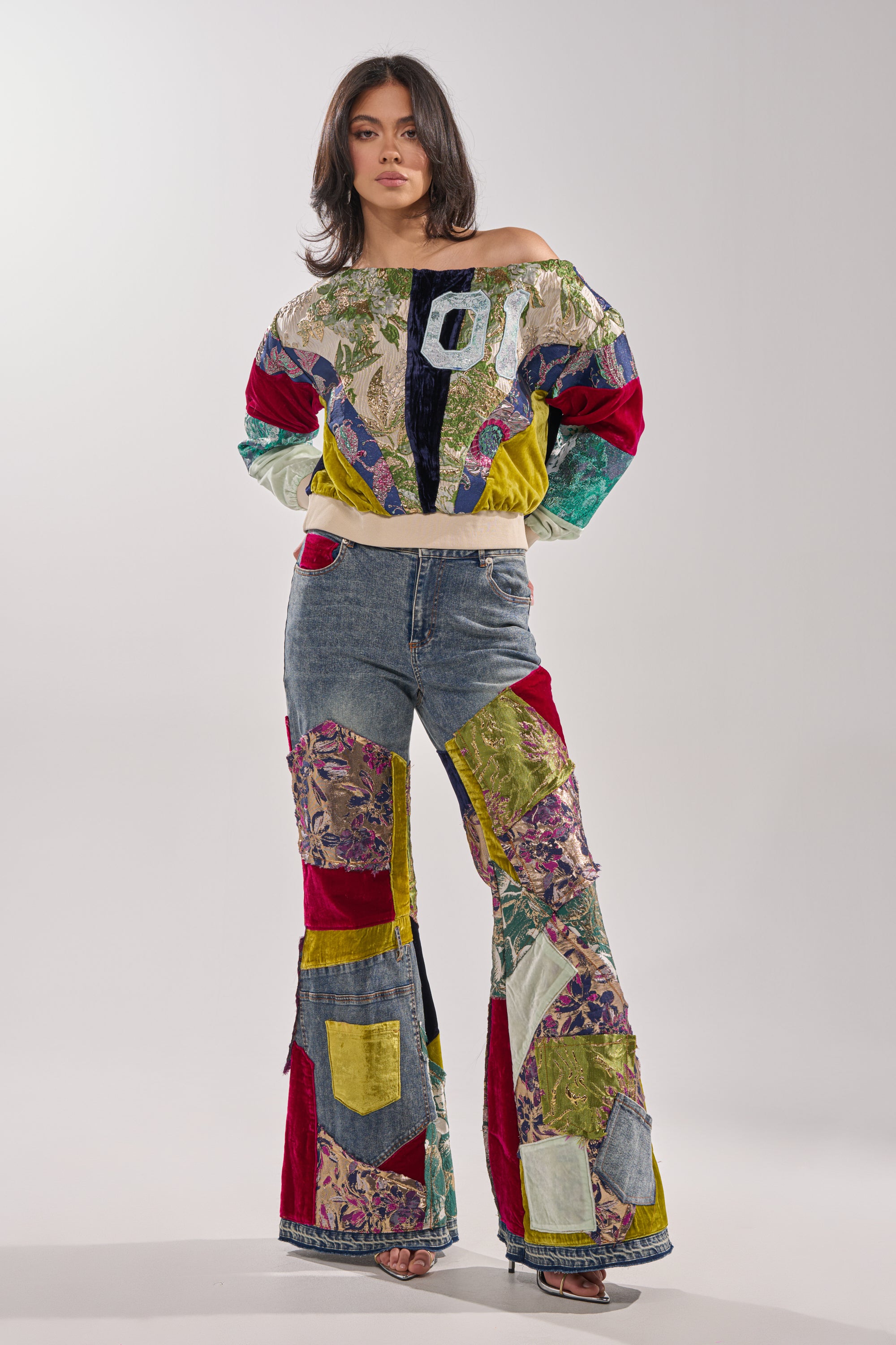 A woman wears the MIXED EMOTION BROCADE PATCHWORK SWEATSHIRT, a colorful off-the-shoulder cropped top featuring various fabric patterns and textures, against a plain light gray background.