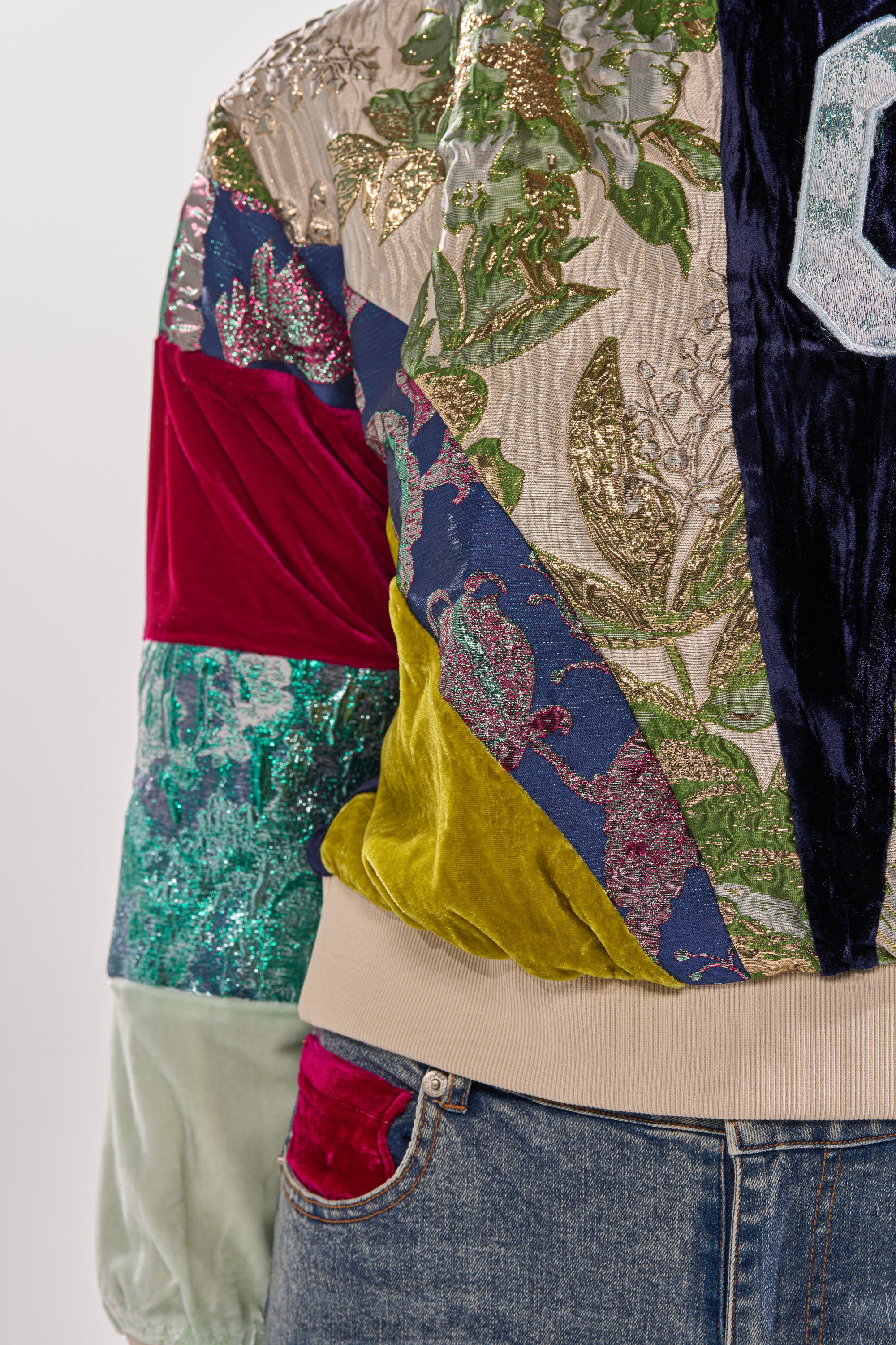 A close-up of the MIXED EMOTION BROCADE PATCHWORK SWEATSHIRT, featuring textured velvet, sequins, and embroidered florals in green, gold, red, blue, and yellow patchwork designs, styled with blue jeans.