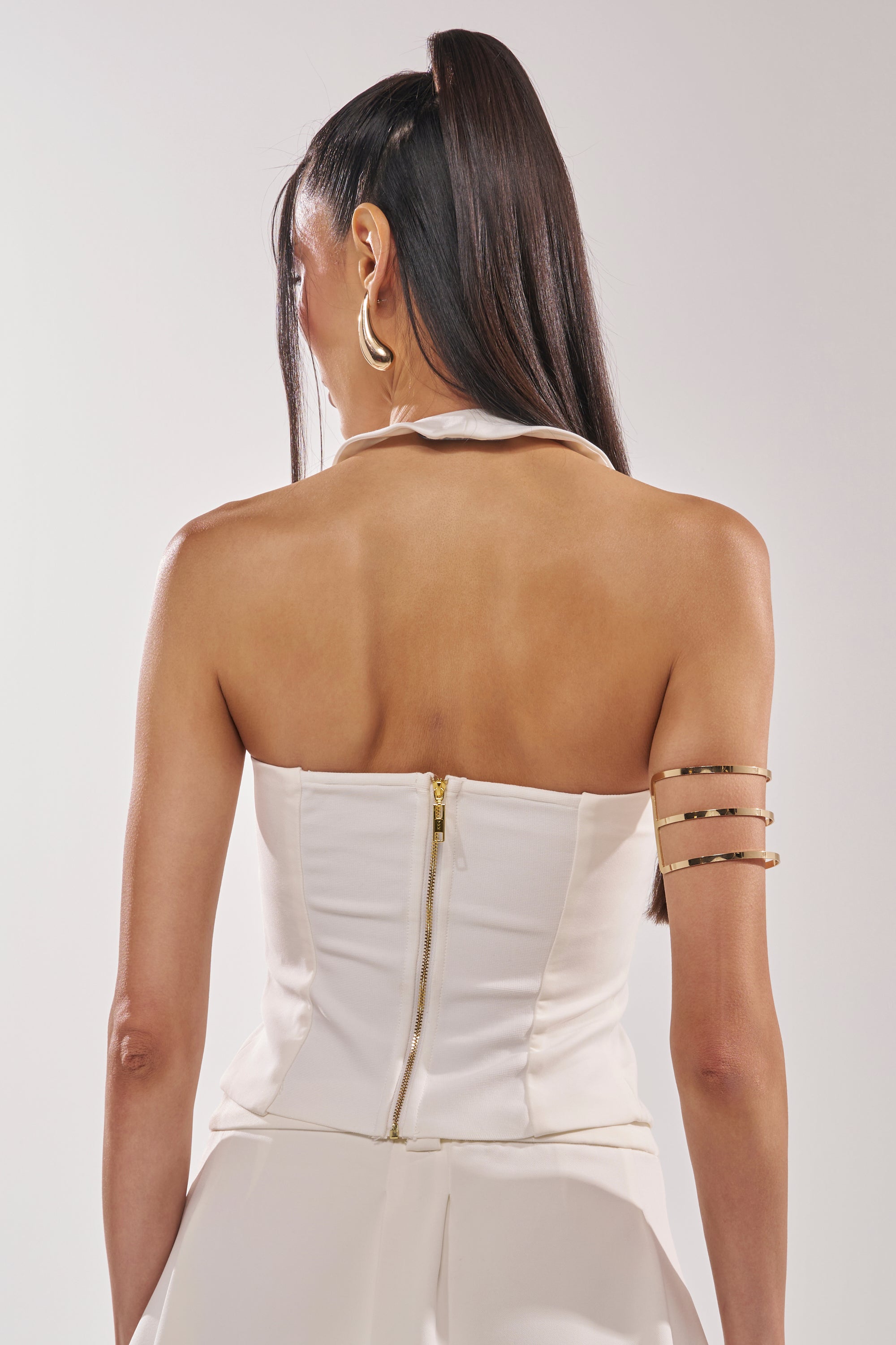 A woman with long, straight hair in a high ponytail is seen from the back, wearing the EYE GOT MY EYES ON YOU PIN DETAIL CORSET TOP with gold accents and accessorizing with large gold hoop earrings for a bold, fashion-forward look.