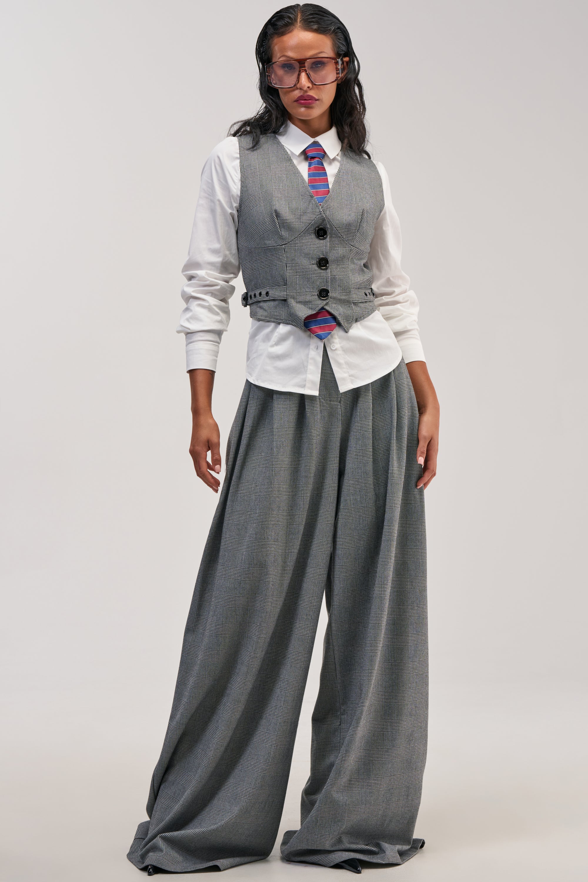 Wearing the WORK HARD PLAY HARD TROUSER—a pair of wide-leg tweed trousers with black buttons—paired with a gray vest over a white shirt and a striped tie, this individual stands against a plain background. Slicked-back hair and large glasses complete the look, exuding classic workwear style.