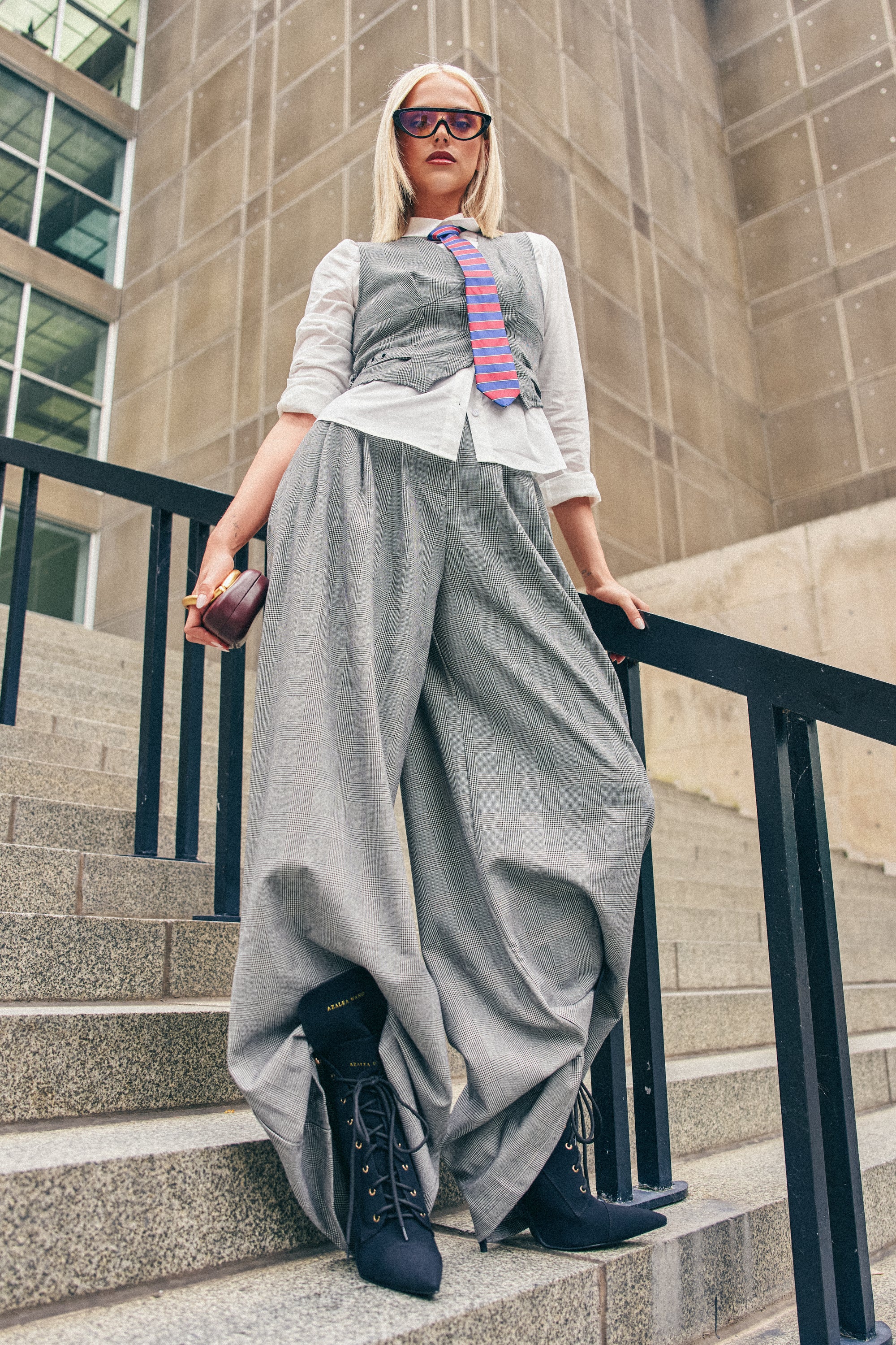 A woman stands on steps in front of a building, striking a confident pose while wearing the WORK HARD PLAY HARD TROUSER—oversized gray wide-leg trousers—paired with a matching vest, white shirt, striped tie, sunglasses, and black boots. She completes her look with a clutch.