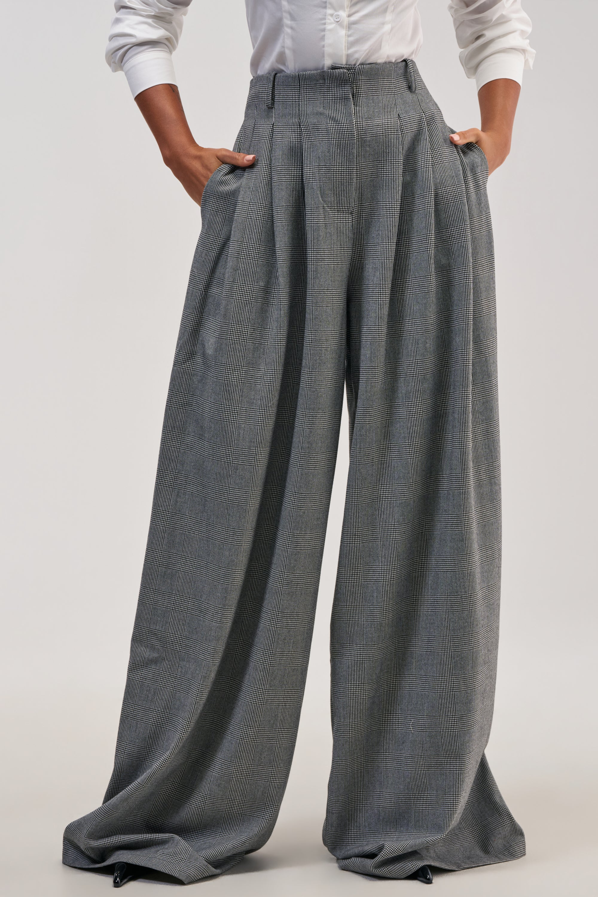 A person wearing a white shirt and the WORK HARD PLAY HARD TROUSER—a pair of high-waisted, pleated, wide-leg trousers that are long and drape to the floor—standing with hands in pockets.