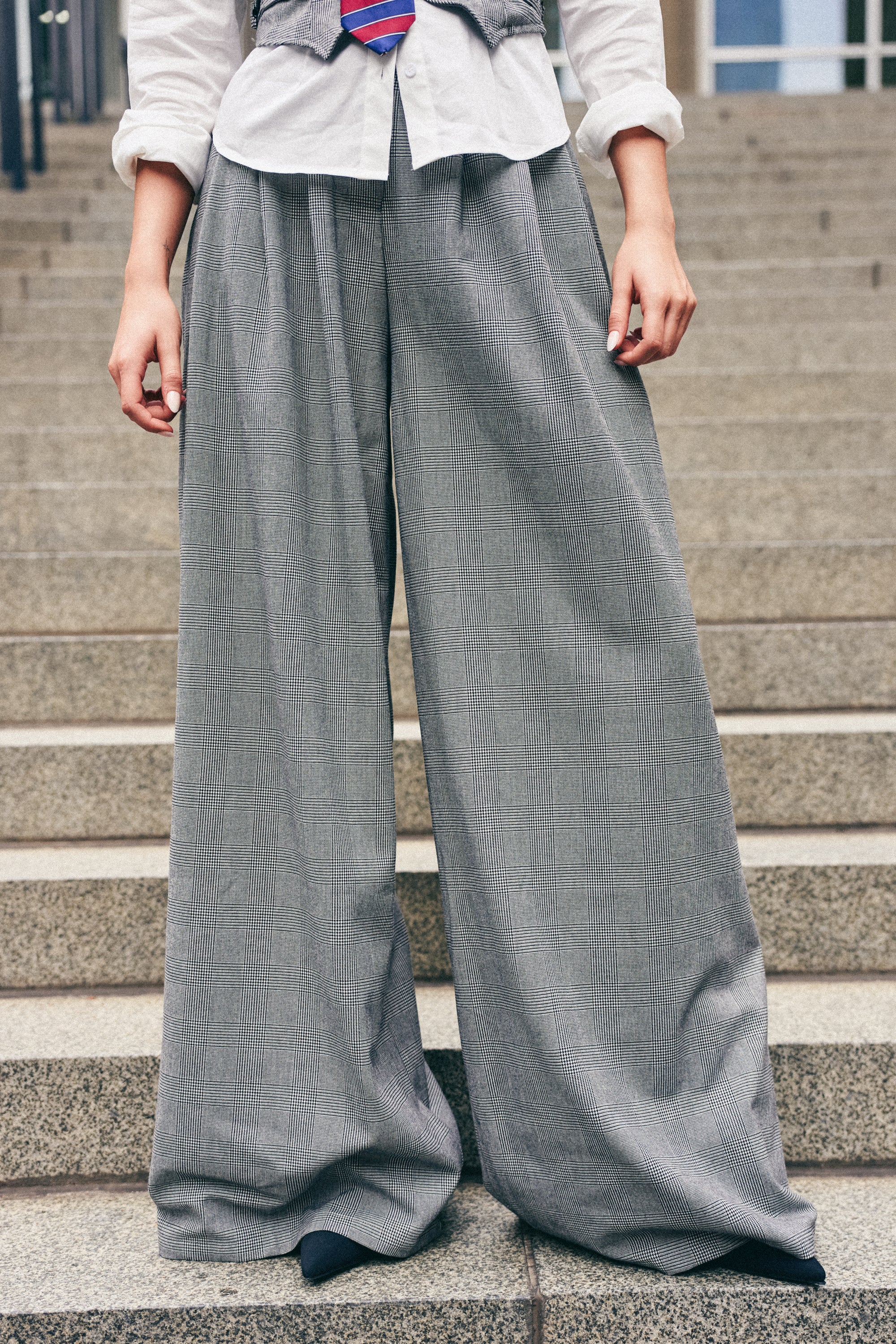 Wearing the WORK HARD PLAY HARD TROUSER in gray, paired with a crisp white shirt and a vest, a person stands on stone steps—only the lower half of their body is visible.
