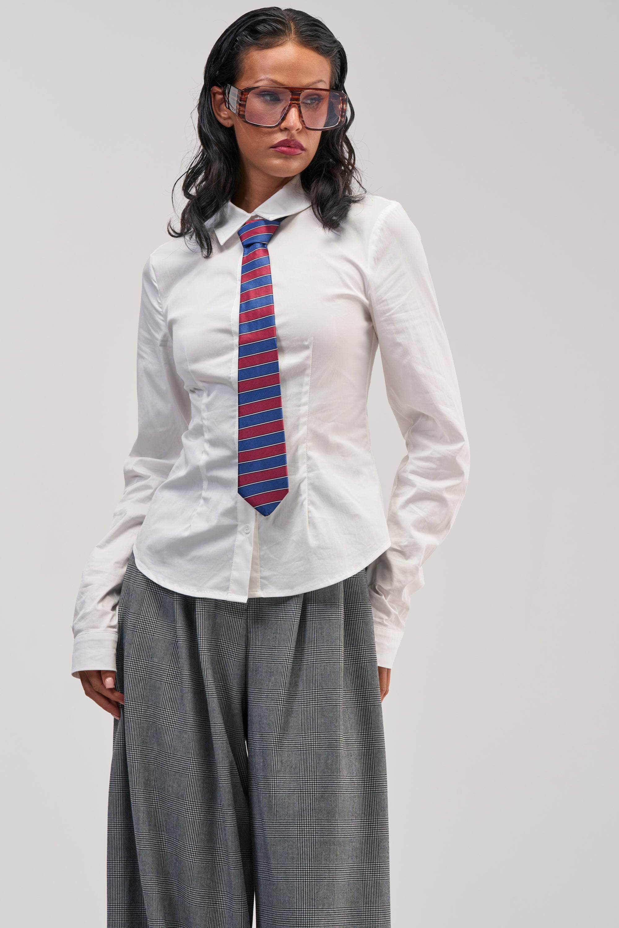 Wearing oversized glasses, loose gray pants, a red and blue striped tie, and the WORK HARD PLAY HARD BUTTON DOWN—a crisp white shirt—this person stands against a plain light background, looking slightly to the side with a neutral expression.