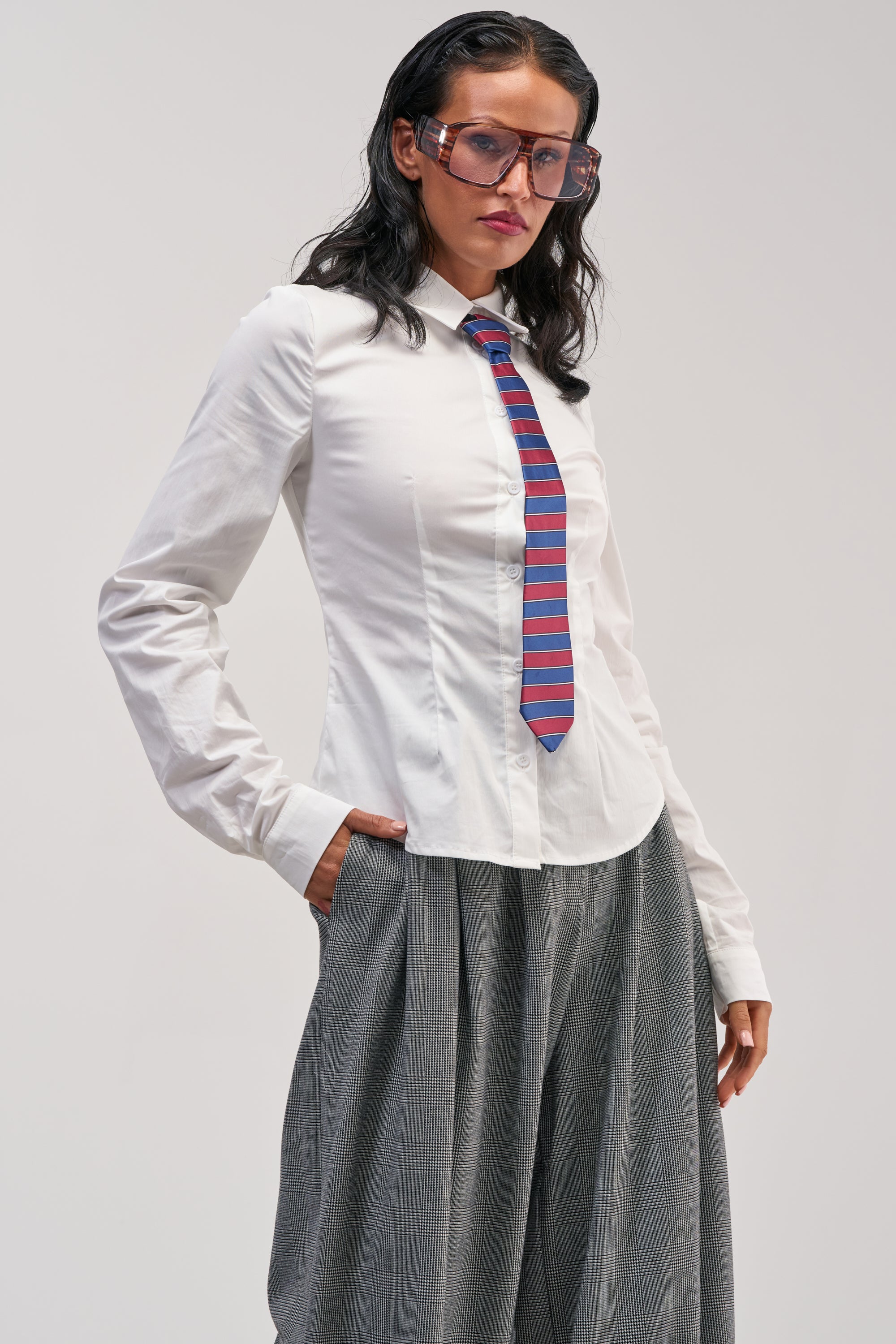Wearing the WORK HARD PLAY HARD BUTTON DOWN, a woman stands with her hands in her pockets against a plain background, styled with a striped tie, gray wide-leg pants, and large sunglasses.