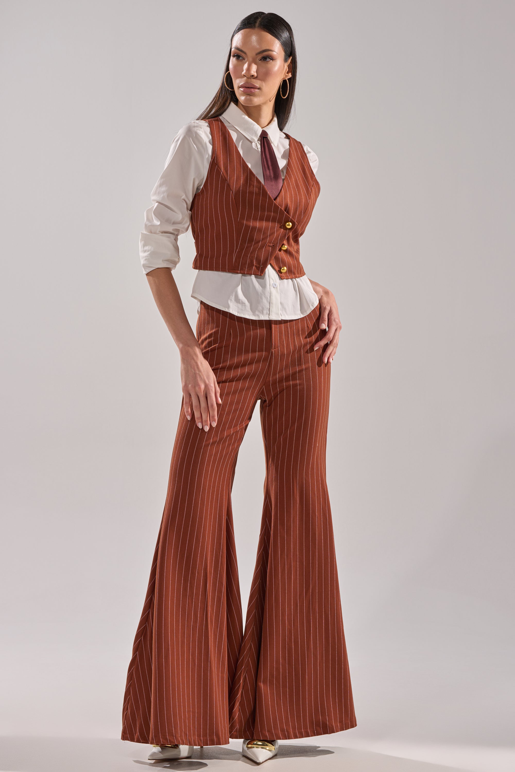 A woman poses confidently in the BUSINESS FIRST STRIPE TROUSER with a matching vest, flared legs, white button-up shirt, and tie. She stands against a plain light background, glancing slightly to the side.