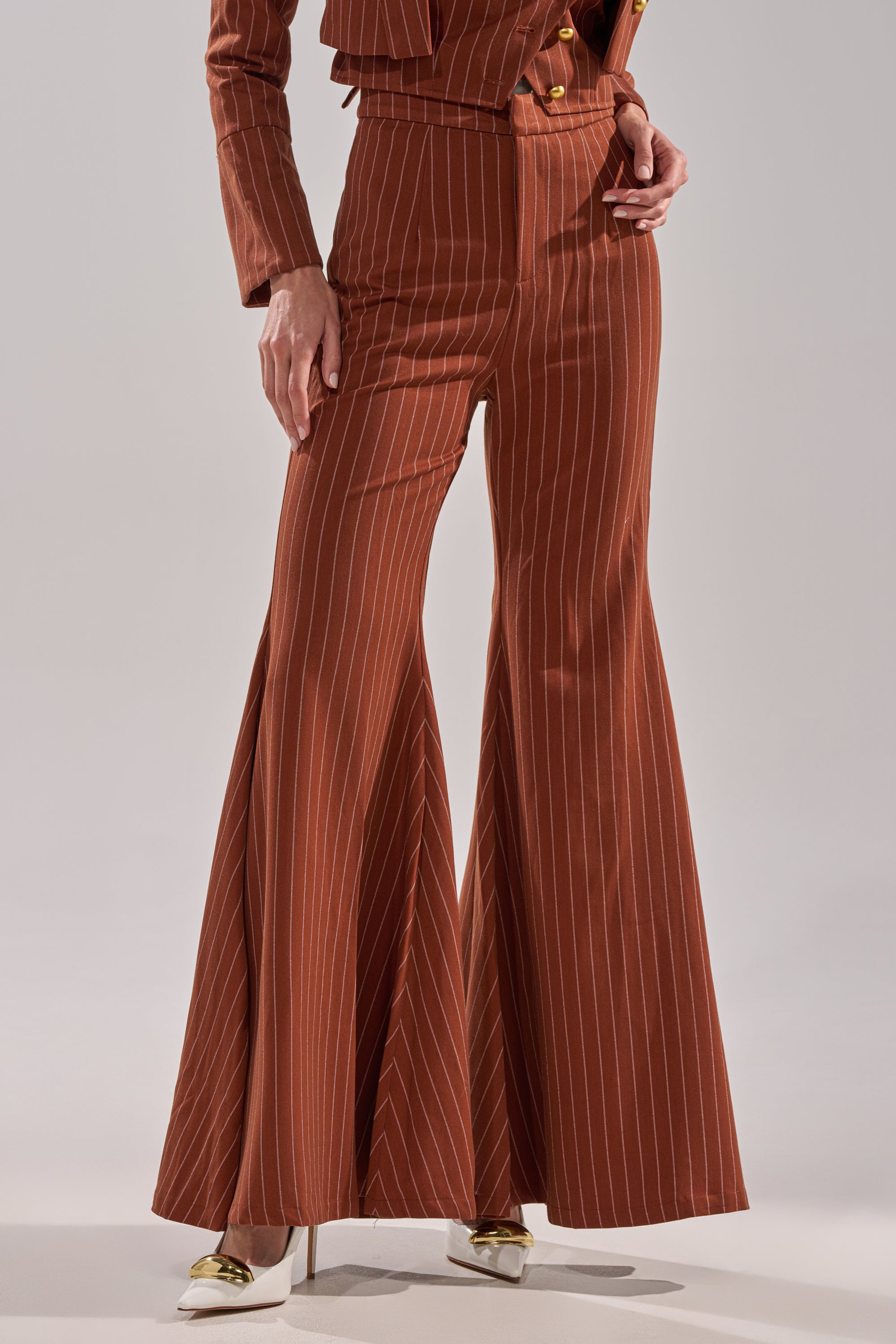 Wearing the BUSINESS FIRST STRIPE TROUSER, a high-rise, brown pinstriped flared-leg style, paired with a matching jacket and white pointed-toe heels; the photo focuses on the lower half of this chic outfit.