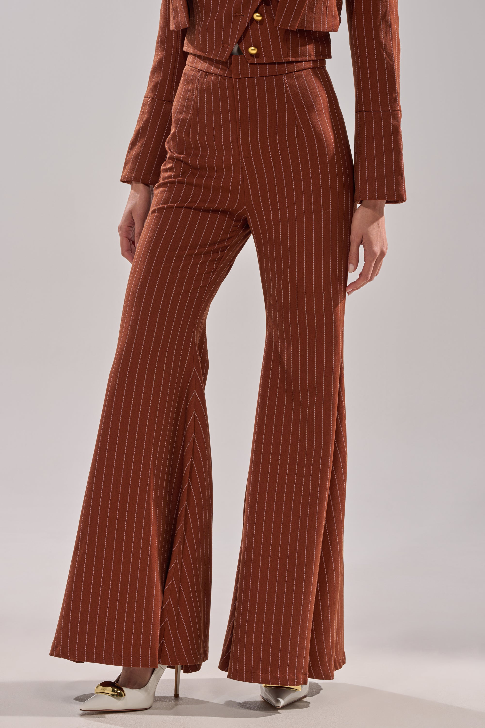 A person wearing the BUSINESS FIRST STRIPE TROUSER—rust-colored, high-rise, wide-leg pinstripe pants—paired with a matching blazer and white pointed-toe heels.