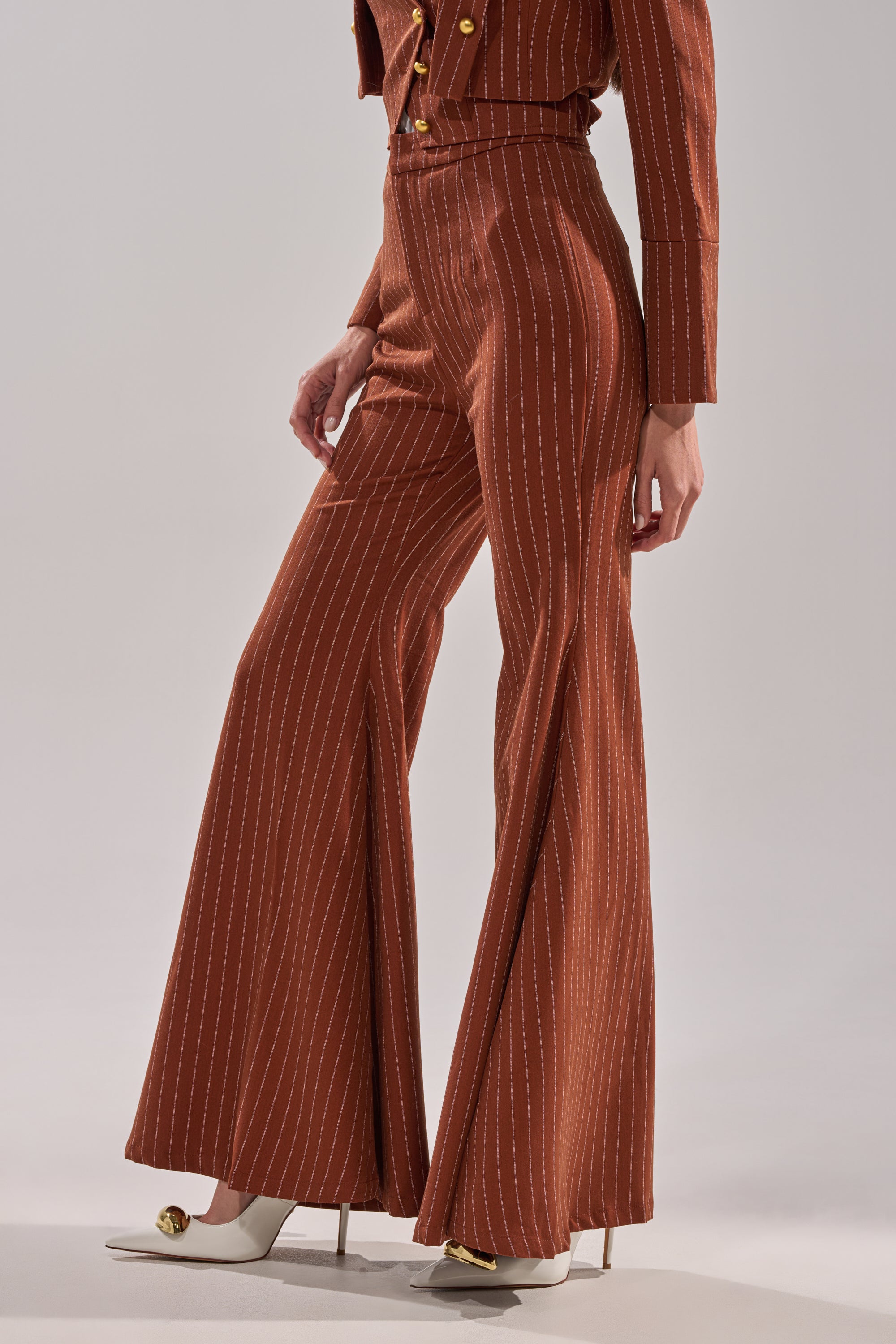 A person stands against a neutral background wearing the BUSINESS FIRST STRIPE TROUSER—high-waisted, brown, striped, flared-leg—and a matching jacket, paired with white pointed-toe heels with gold accents.