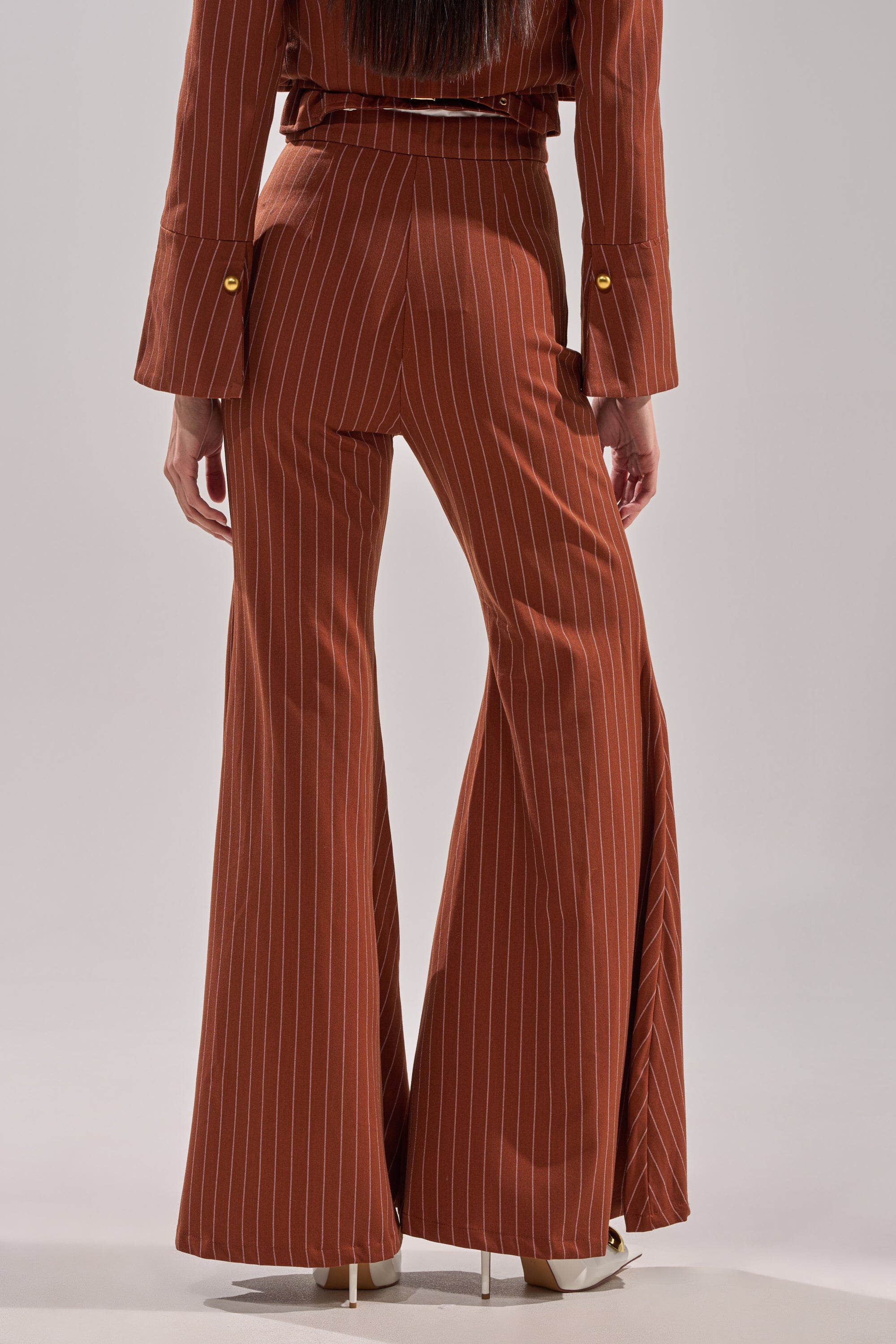 Seen from the back, a person wears the BUSINESS FIRST STRIPE TROUSER in a high-rise, wide-leg fit with brown stripes and a matching jacket, styled with white heels to emphasize the flared silhouette.
