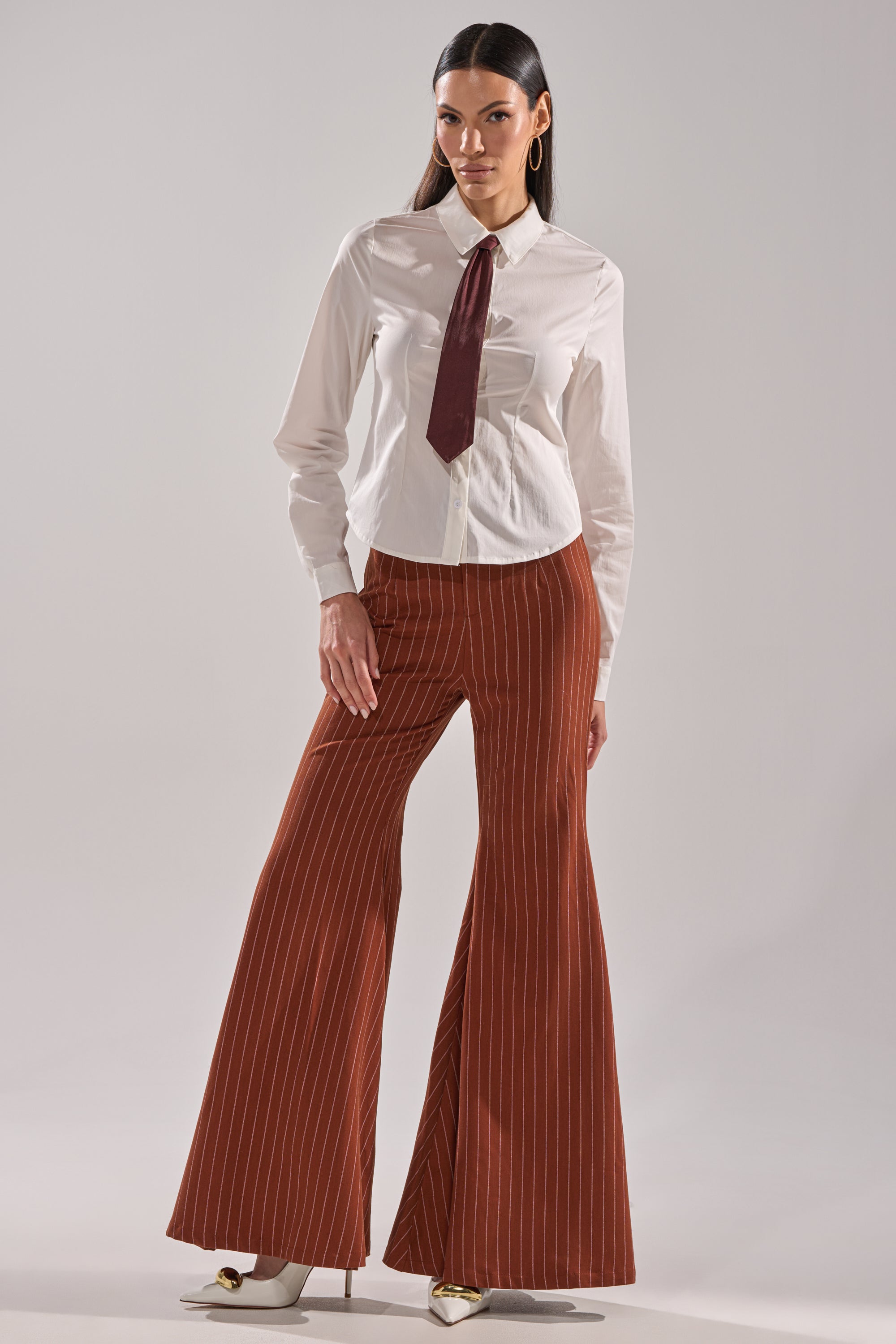 A woman poses confidently in the BUSINESS FIRST BUTTON DOWN, a tailored white shirt with a detachable satin necktie, paired with high-waisted brown pinstriped flared pants. She has long dark hair and stands against a plain background.