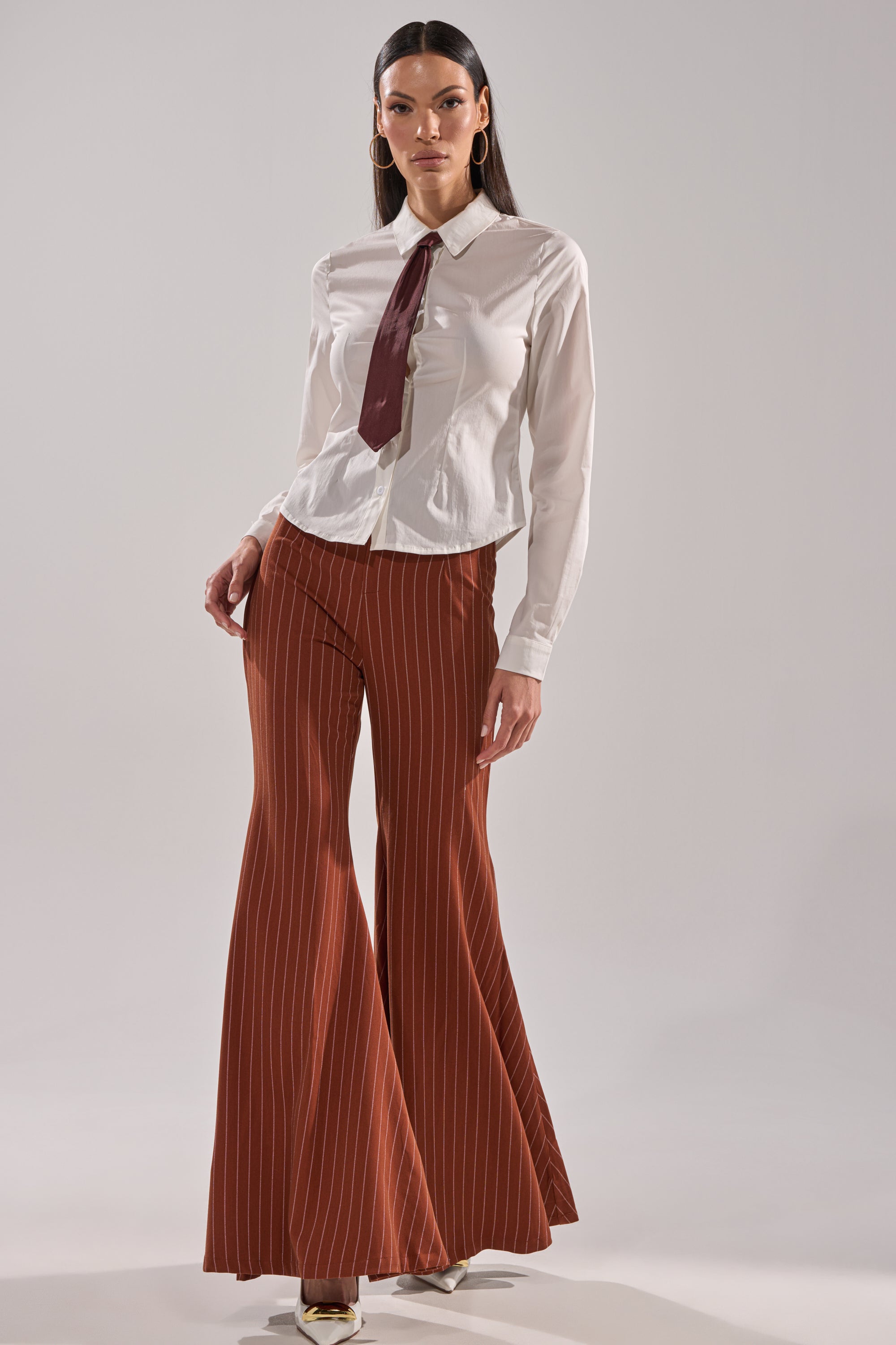 A woman wears the BUSINESS FIRST BUTTON DOWN blouse with a detachable satin necktie, styled with high-waisted rust pinstripe flared pants and pointed-toe shoes, posing against a plain light background.