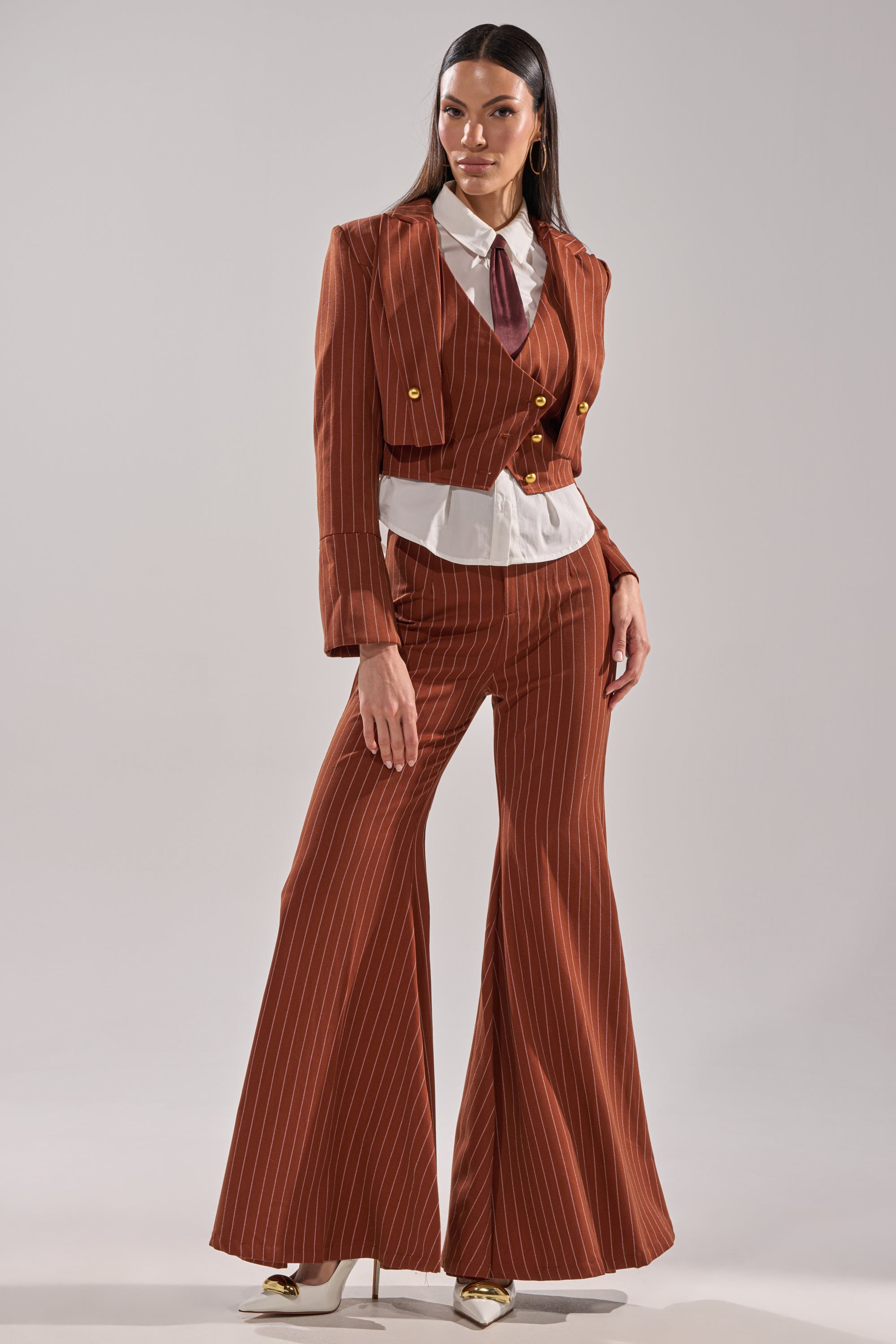 A woman stands confidently in the BUSINESS FIRST BUTTON DOWN, styled with brown pinstripe flared trousers, a cropped double-breasted blazer, detachable satin necktie, and gold heels against a plain light background.