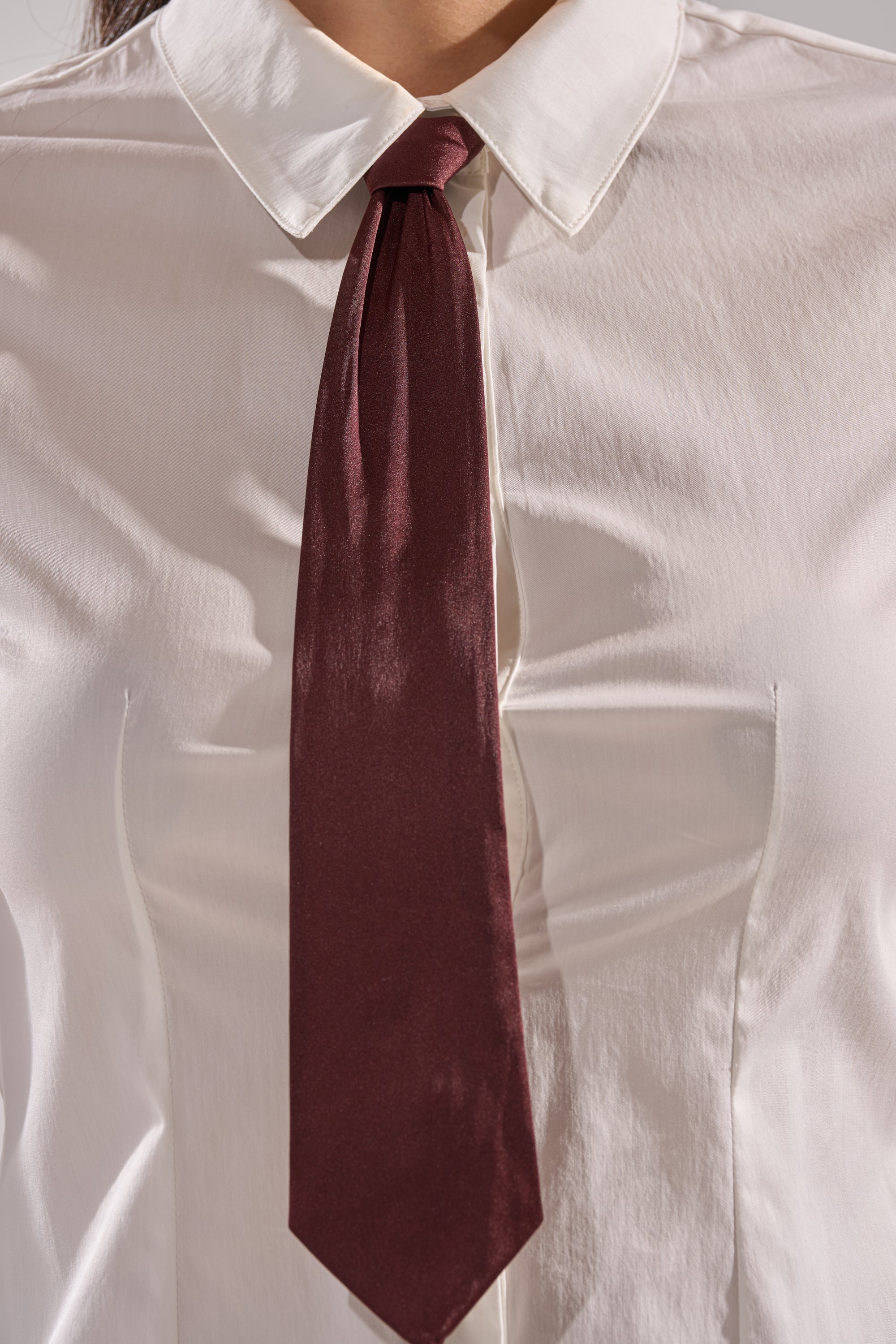 A person models the BUSINESS FIRST BUTTON DOWN, a tailored white blouse styled as a button down shirt with a dark maroon tie, shown from shoulders to chest.