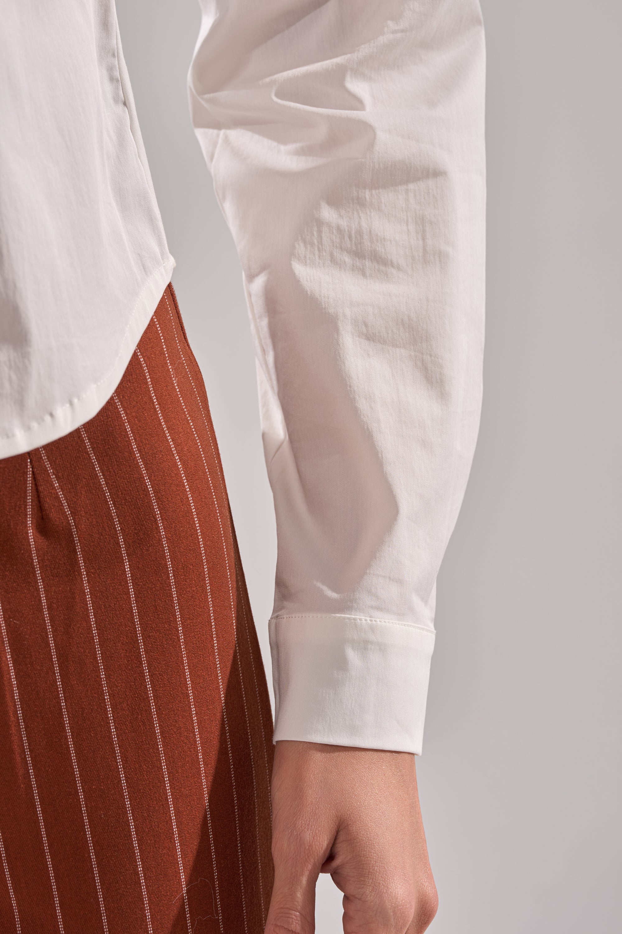 A person is shown from the waist down wearing the BUSINESS FIRST BUTTON DOWN in white, paired with brown pants featuring white vertical pinstripes, with their arm relaxed at their side.