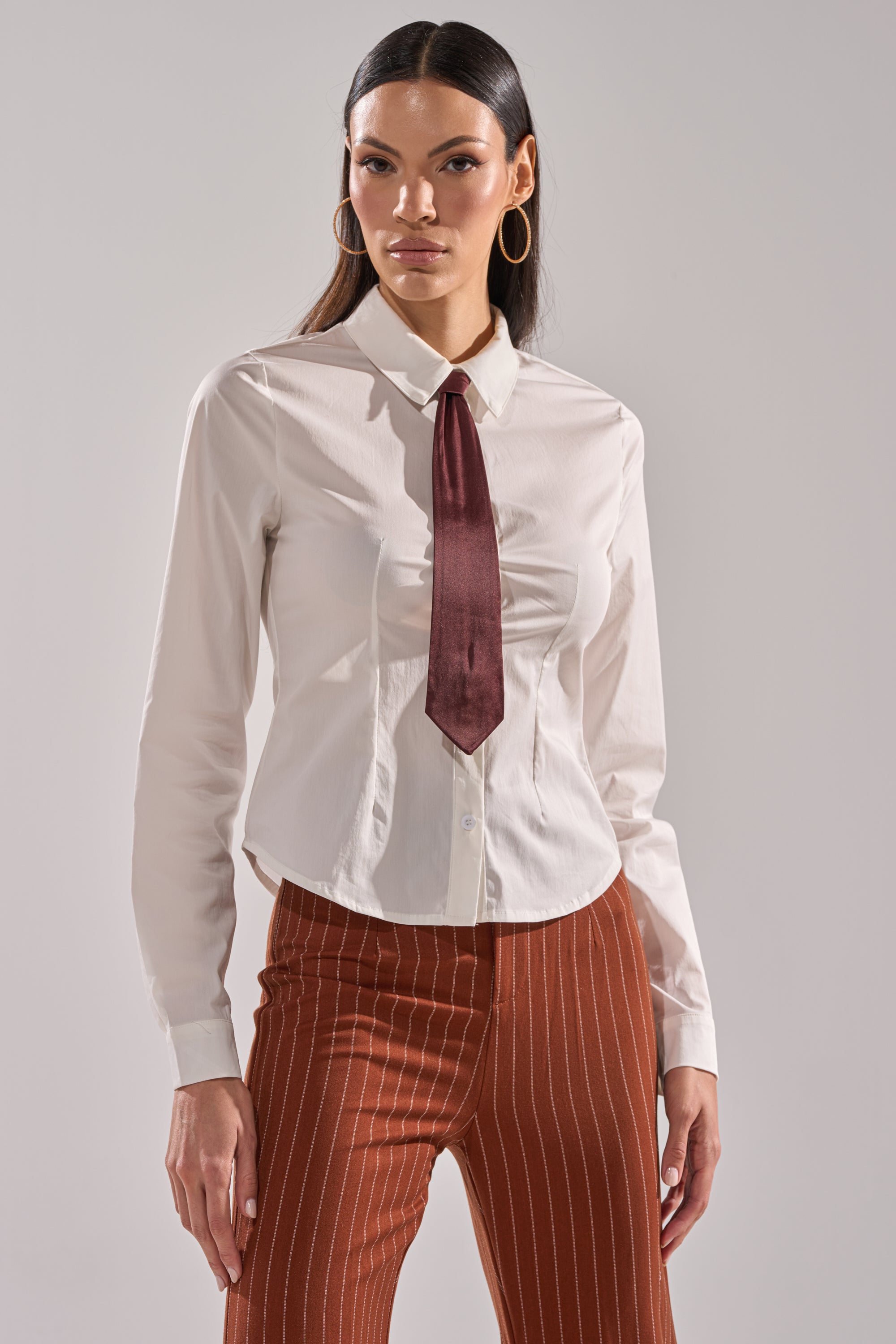 A woman with long dark hair and hoop earrings faces forward in the BUSINESS FIRST BUTTON DOWN, styled with a dark red satin necktie and rust-colored pinstriped pants, standing against a plain background with a neutral expression.