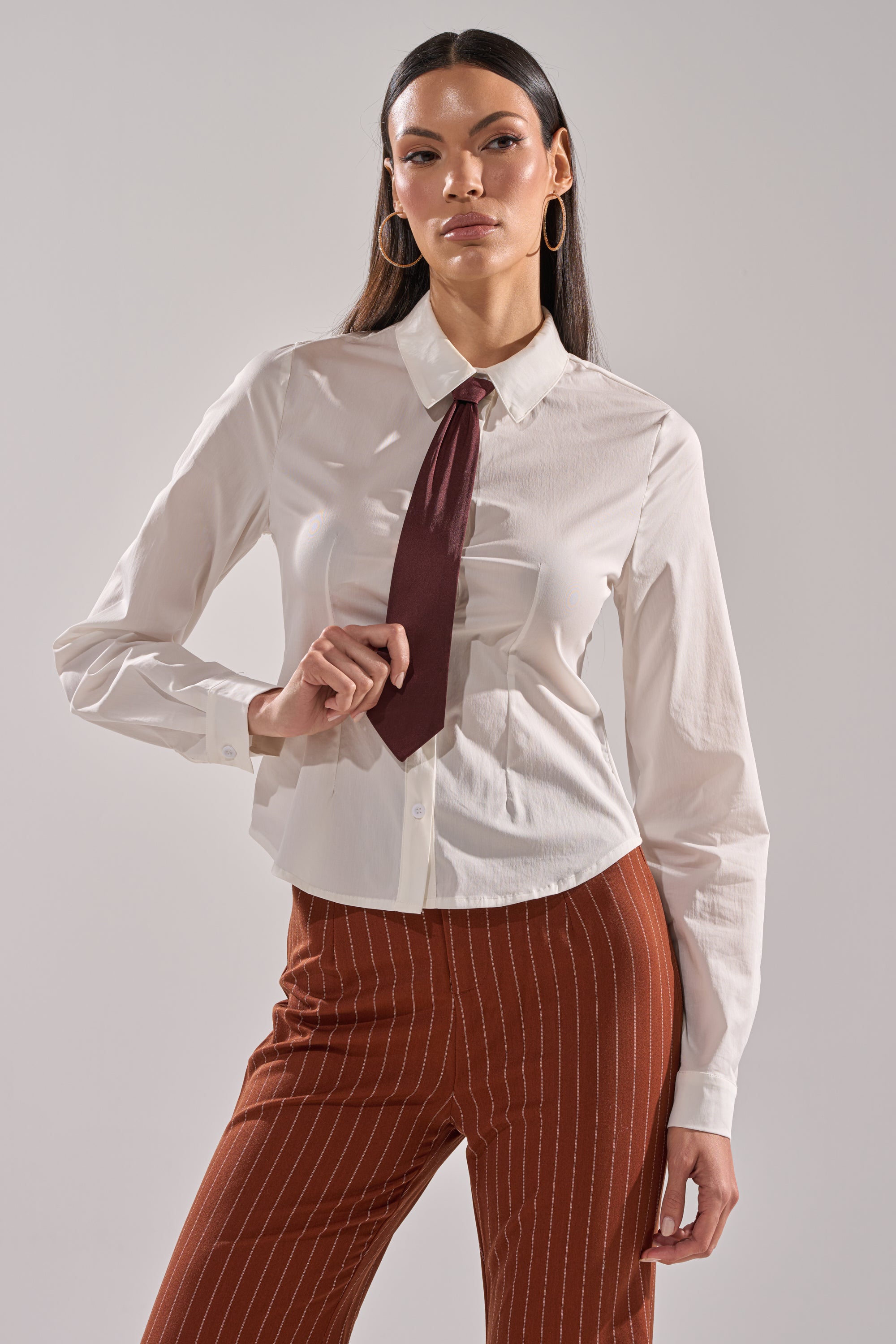 A woman wearing the BUSINESS FIRST BUTTON DOWN, styled with a detachable satin necktie and brown pinstripe pants, stands confidently against a plain background, looking directly at the camera.