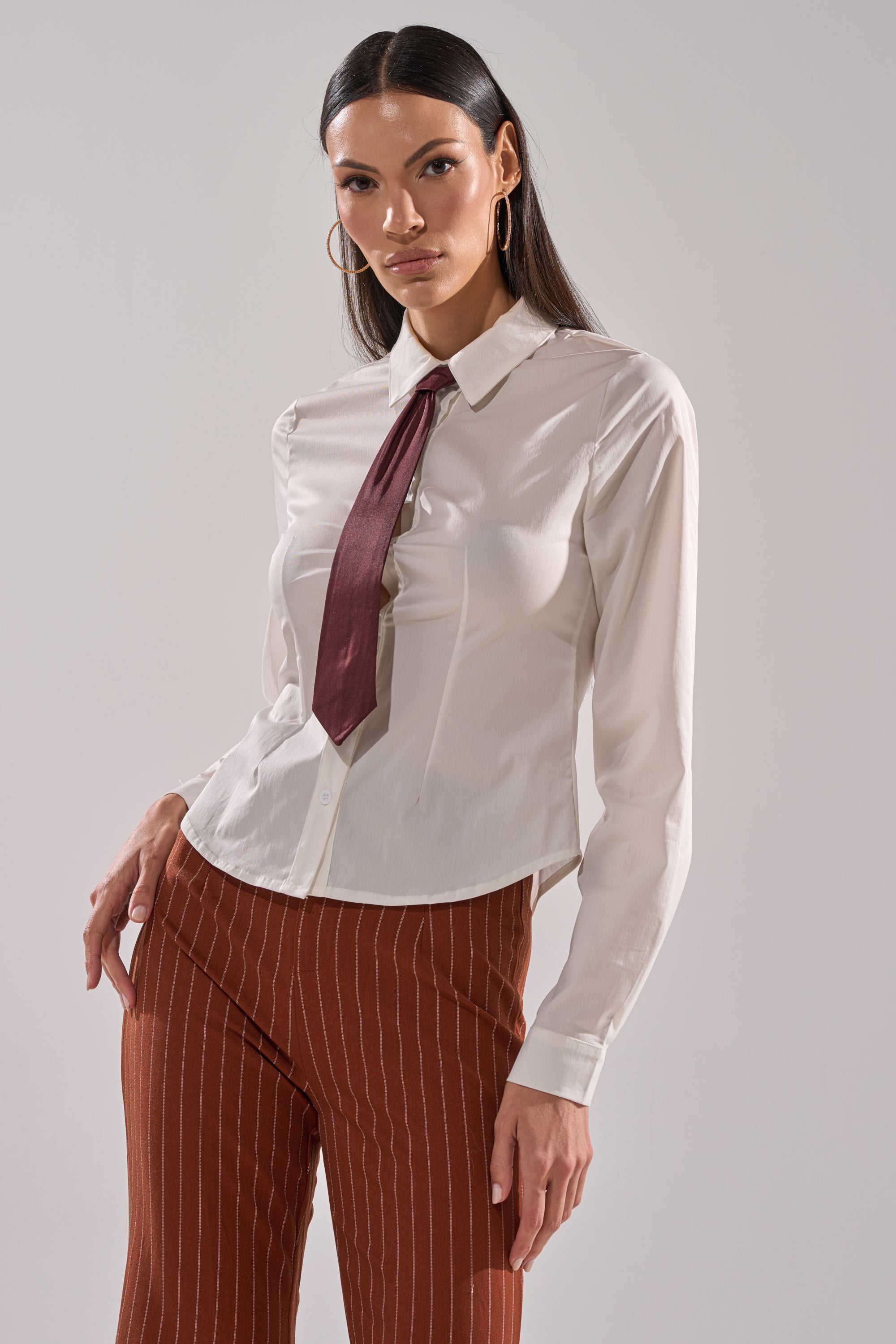 A woman stands confidently against a light gray background wearing the BUSINESS FIRST BUTTON DOWN blouse with a detachable satin necktie and high-waisted brown pants featuring white vertical stripes.