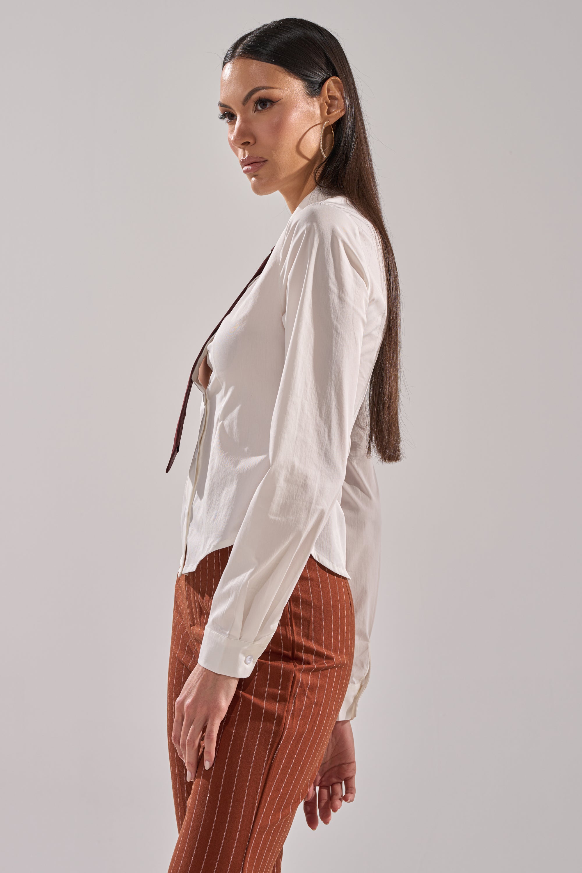 A woman with long dark hair wears the BUSINESS FIRST BUTTON DOWN and brown pinstripe pants, standing in profile against a plain light background.