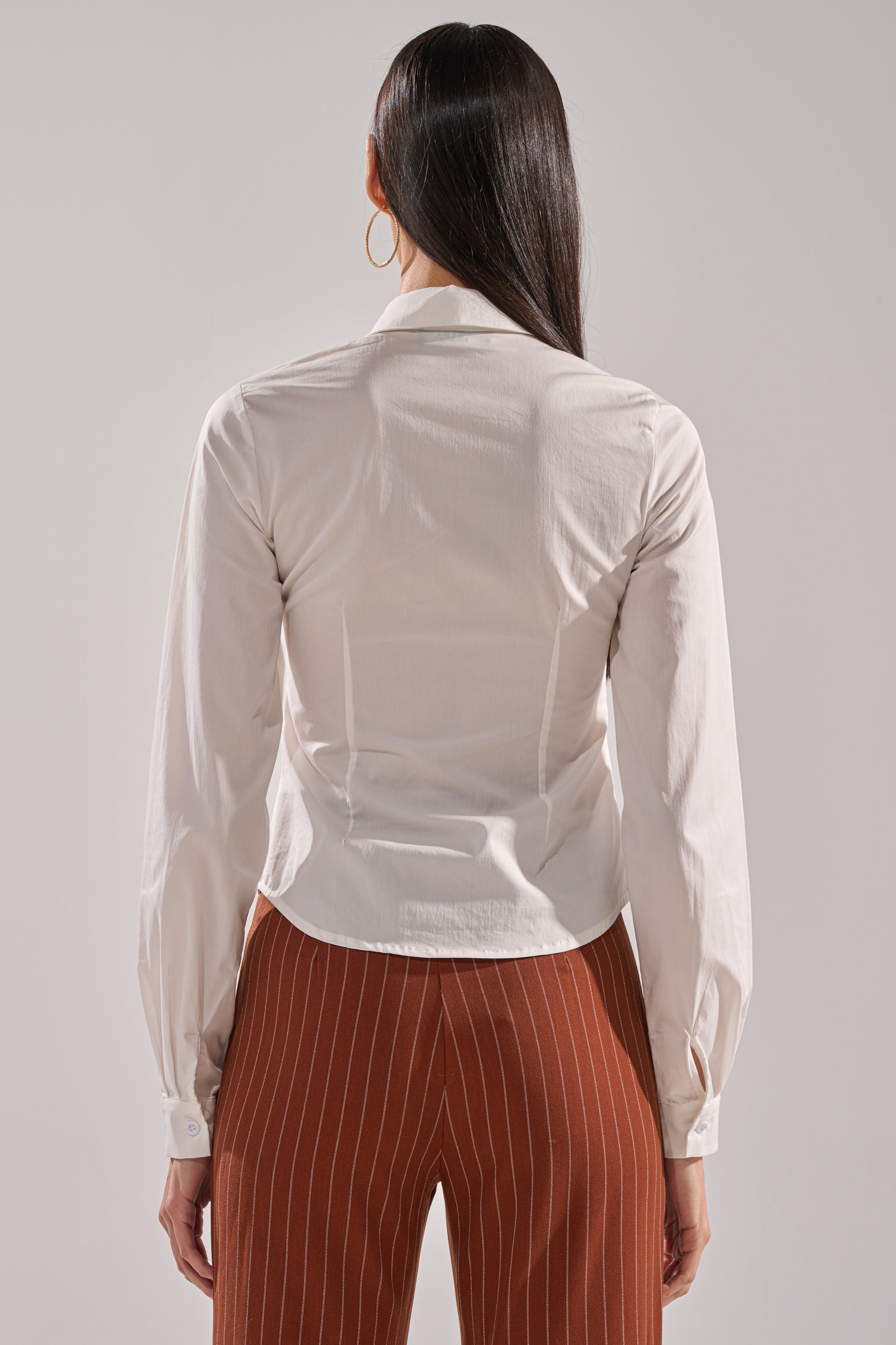 A person with long dark hair, seen from behind, is wearing the BUSINESS FIRST BUTTON DOWN in white and brown pants with white vertical stripes against a plain light-colored background.