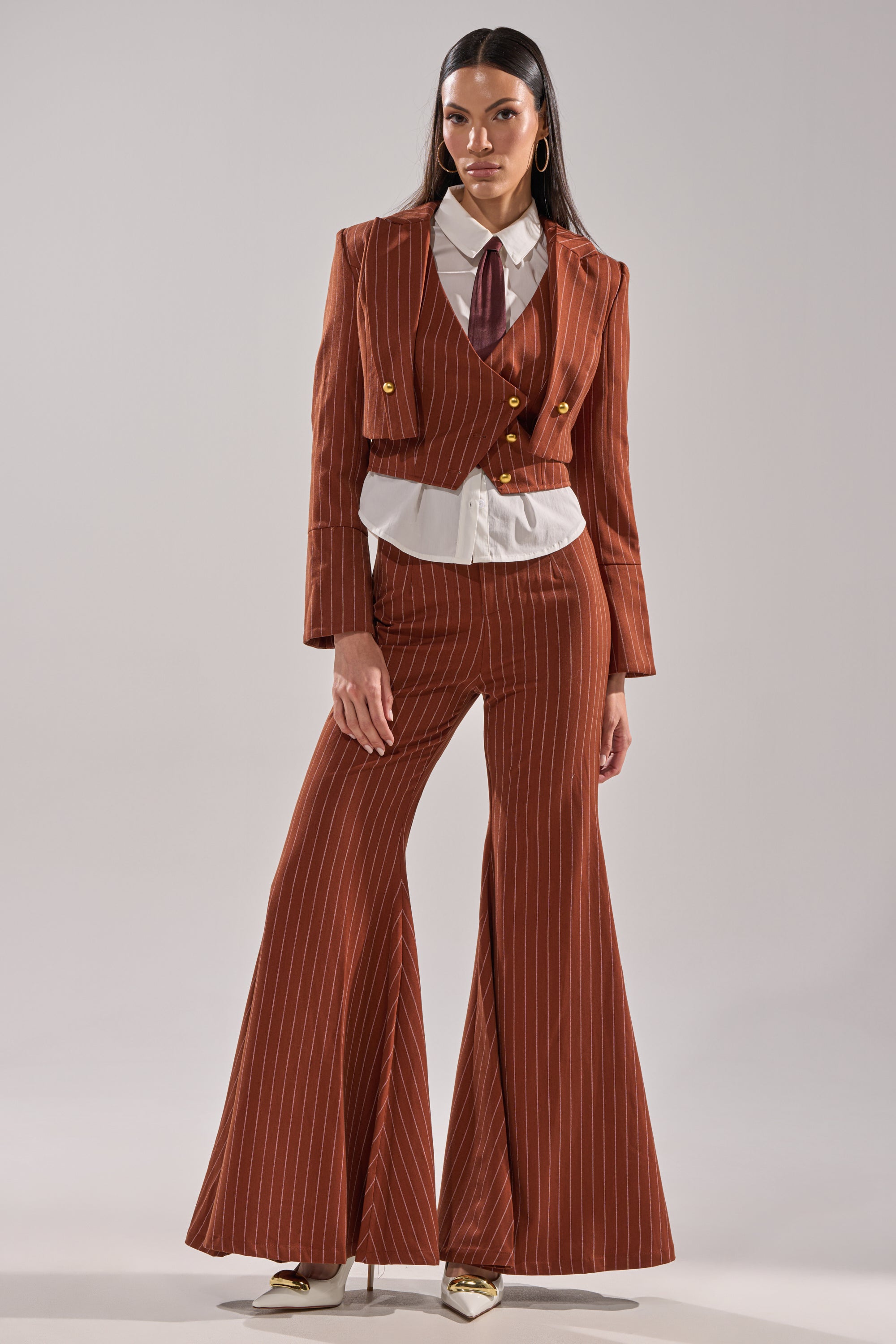 A woman stands confidently in the BUSINESS FIRST STRIPE BLAZER—a rust-colored cropped pinstripe blazer—styled with matching flared trousers, a white shirt, burgundy tie, and gold heels against a plain background.