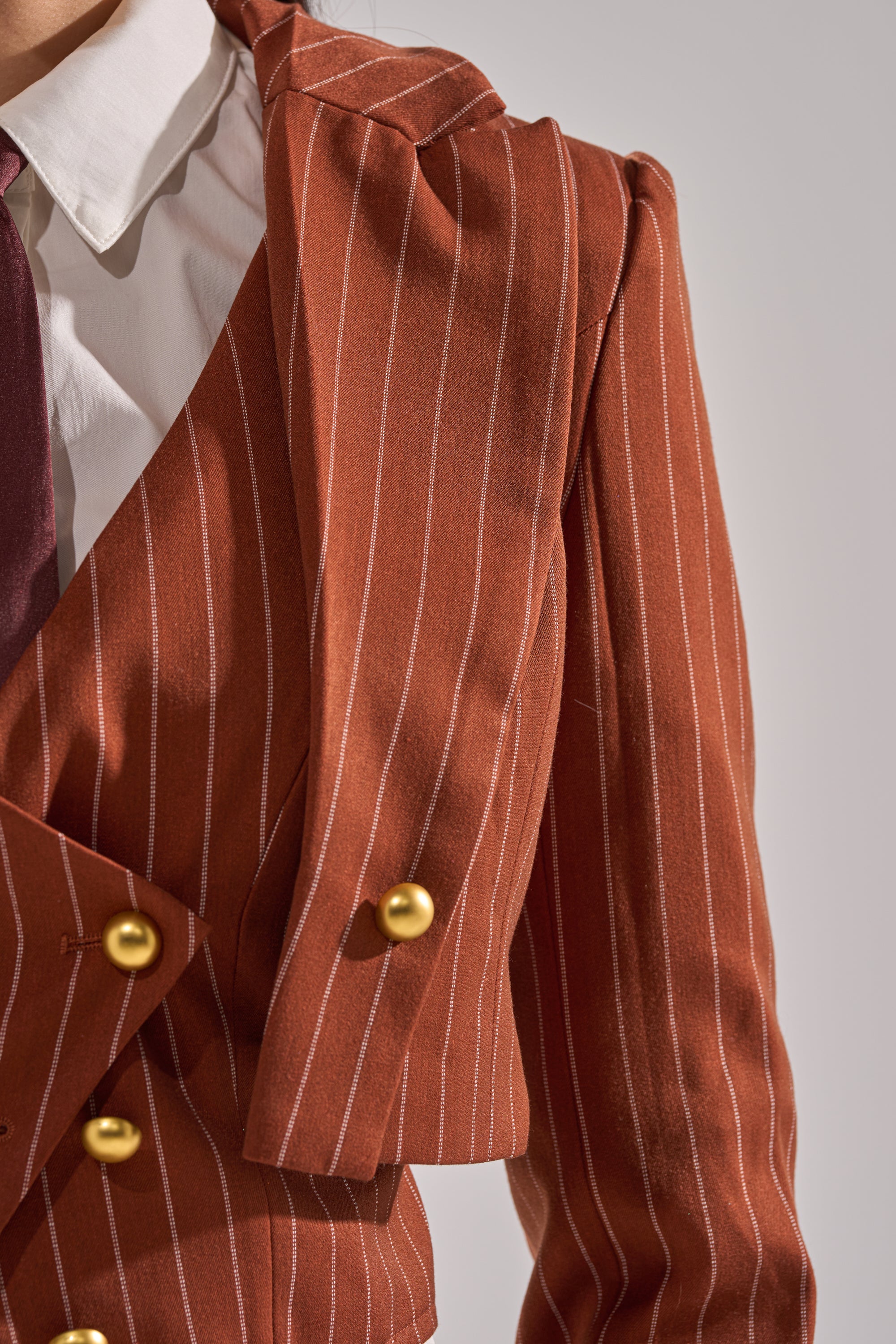 A close-up shows a person wearing the BUSINESS FIRST STRIPE BLAZER, a brown pinstripe blazer with gold buttons, featuring an asymmetrical draped shoulder, styled with a white dress shirt and burgundy tie.
