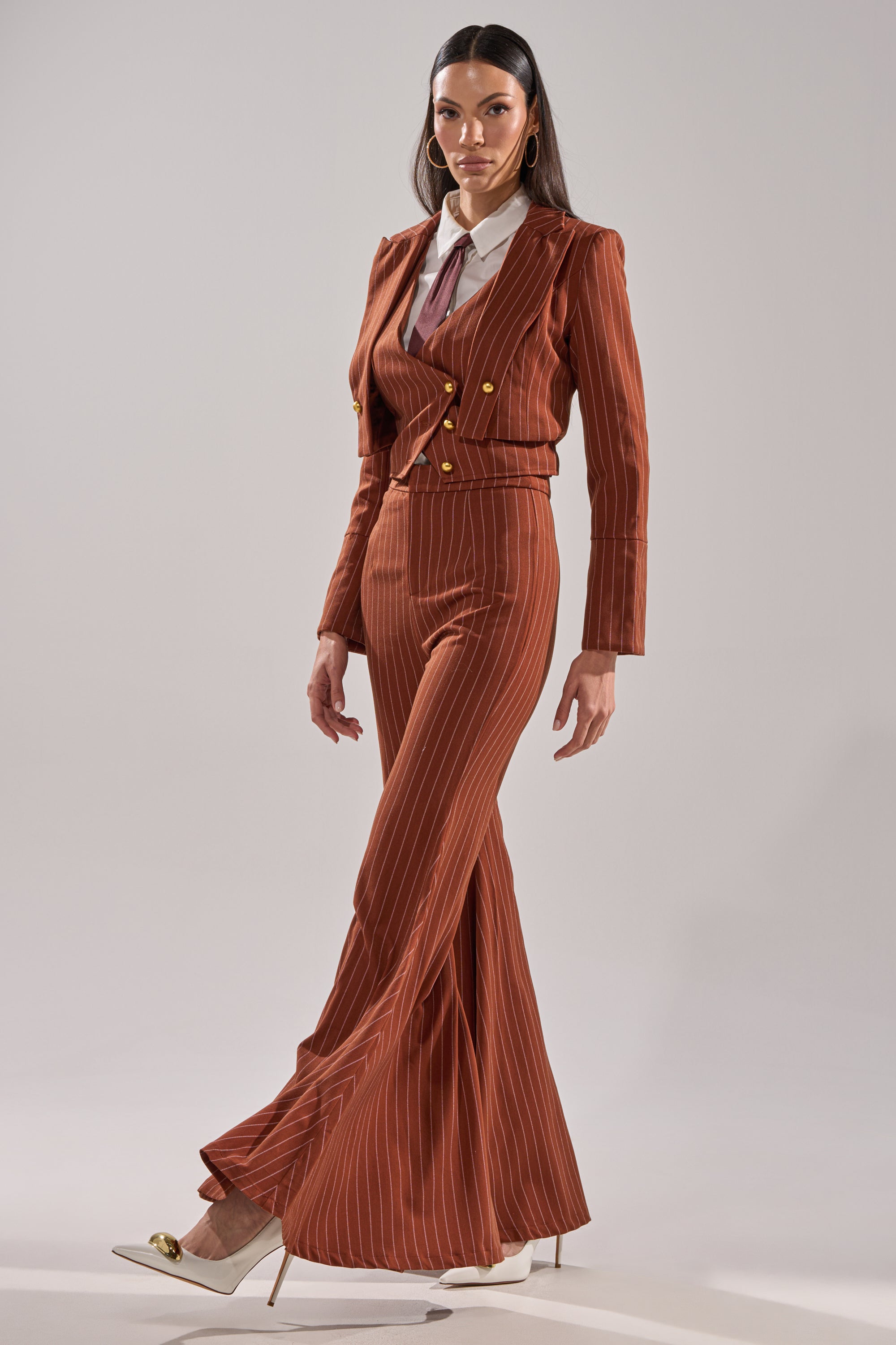 A woman stands confidently in the BUSINESS FIRST STRIPE BLAZER, a rust-colored pinstripe jacket with gold buttons and wide-leg pants, styled with a white shirt, maroon tie, and cream heels against a neutral background.