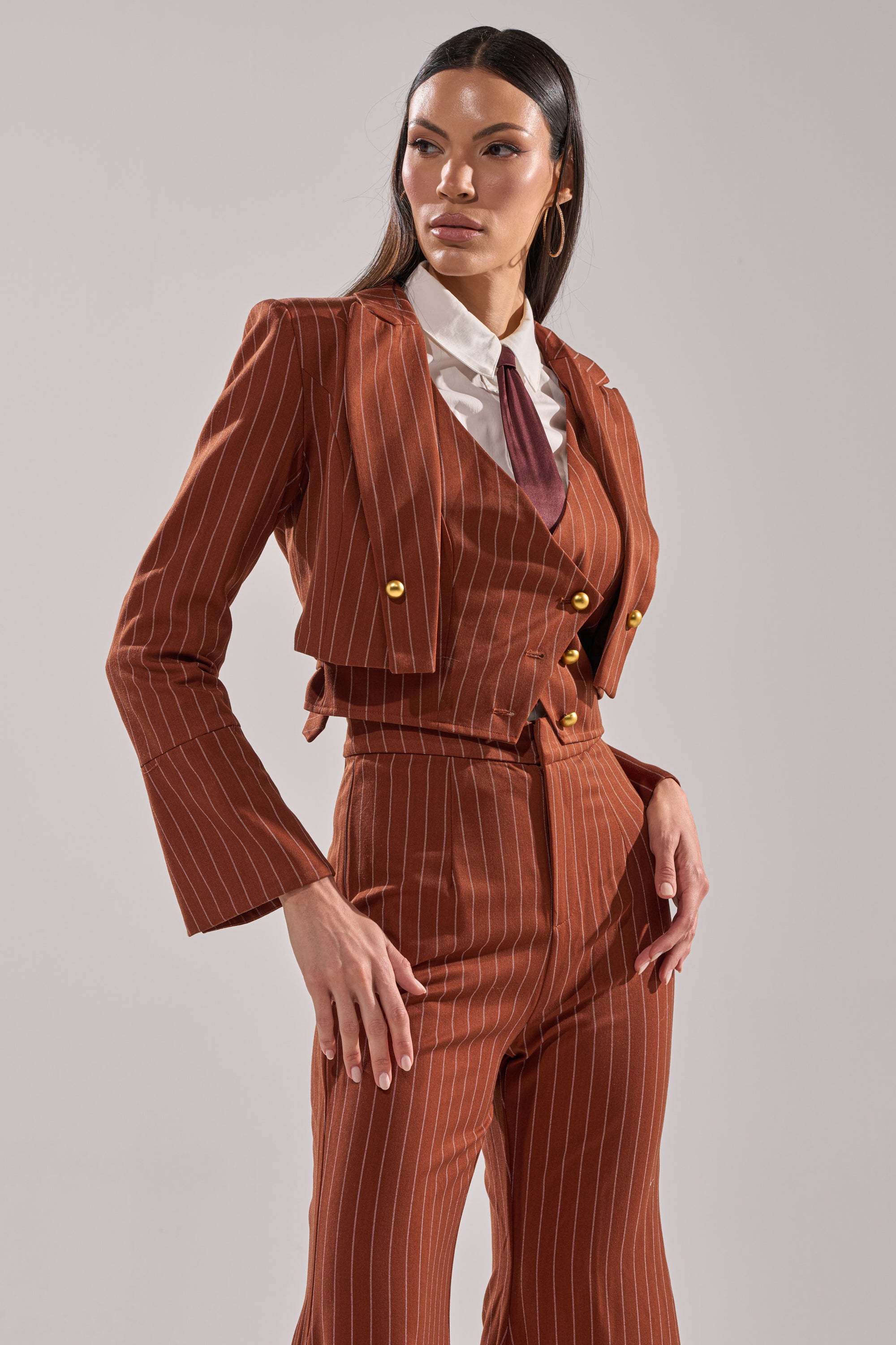 A woman exudes confidence in the BUSINESS FIRST STRIPE BLAZER, styled with wide-legged pants, a cropped cut, a white collared shirt, and maroon tie against a plain background.
