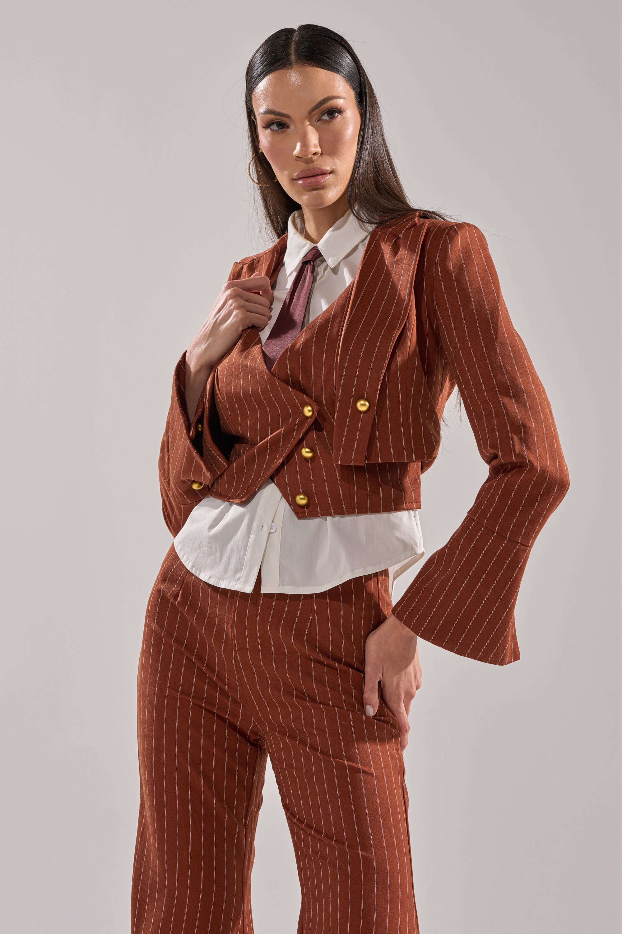 A woman poses confidently in the BUSINESS FIRST STRIPE BLAZER with flared sleeves and matching pants over a white shirt and red tie, set against a plain light background.