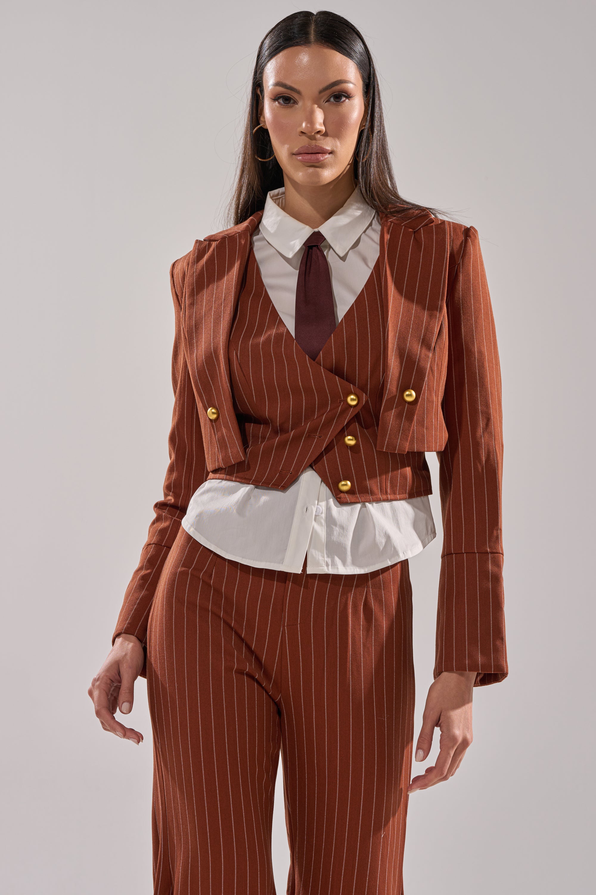 A woman poses confidently against a light gray background wearing the BUSINESS FIRST STRIPE BLAZER—rust-colored with gold buttons—paired with a white collared shirt and matching tie.
