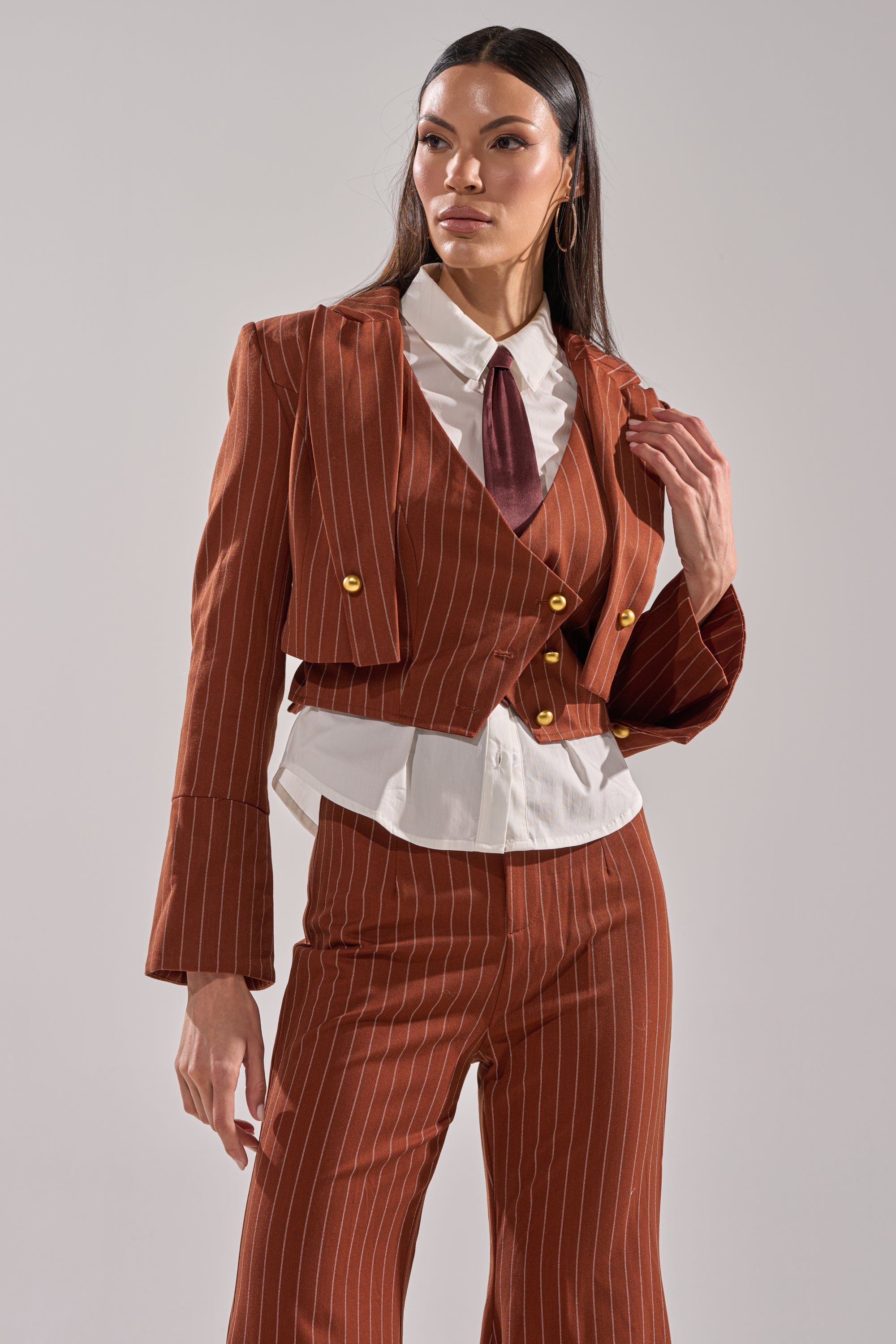 A woman in the BUSINESS FIRST STRIPE BLAZER—a brown pinstripe jacket with gold buttons—pairs it with a white collared shirt and maroon tie, standing against a plain background and looking confidently to the side.