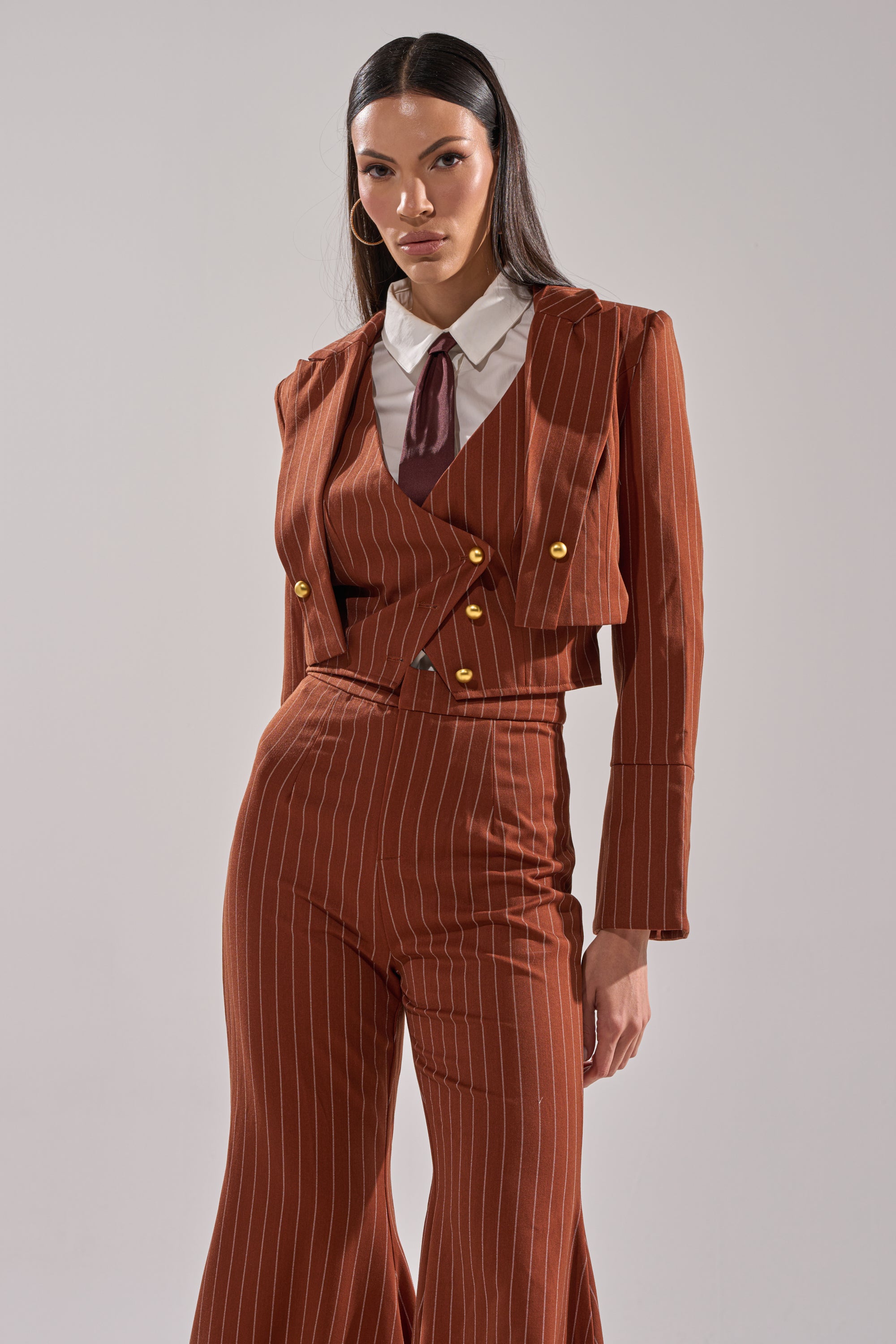 A woman with long dark hair wears the BUSINESS FIRST STRIPE BLAZER with gold buttons, styled over a white collared shirt and burgundy tie, standing confidently against a plain light background.