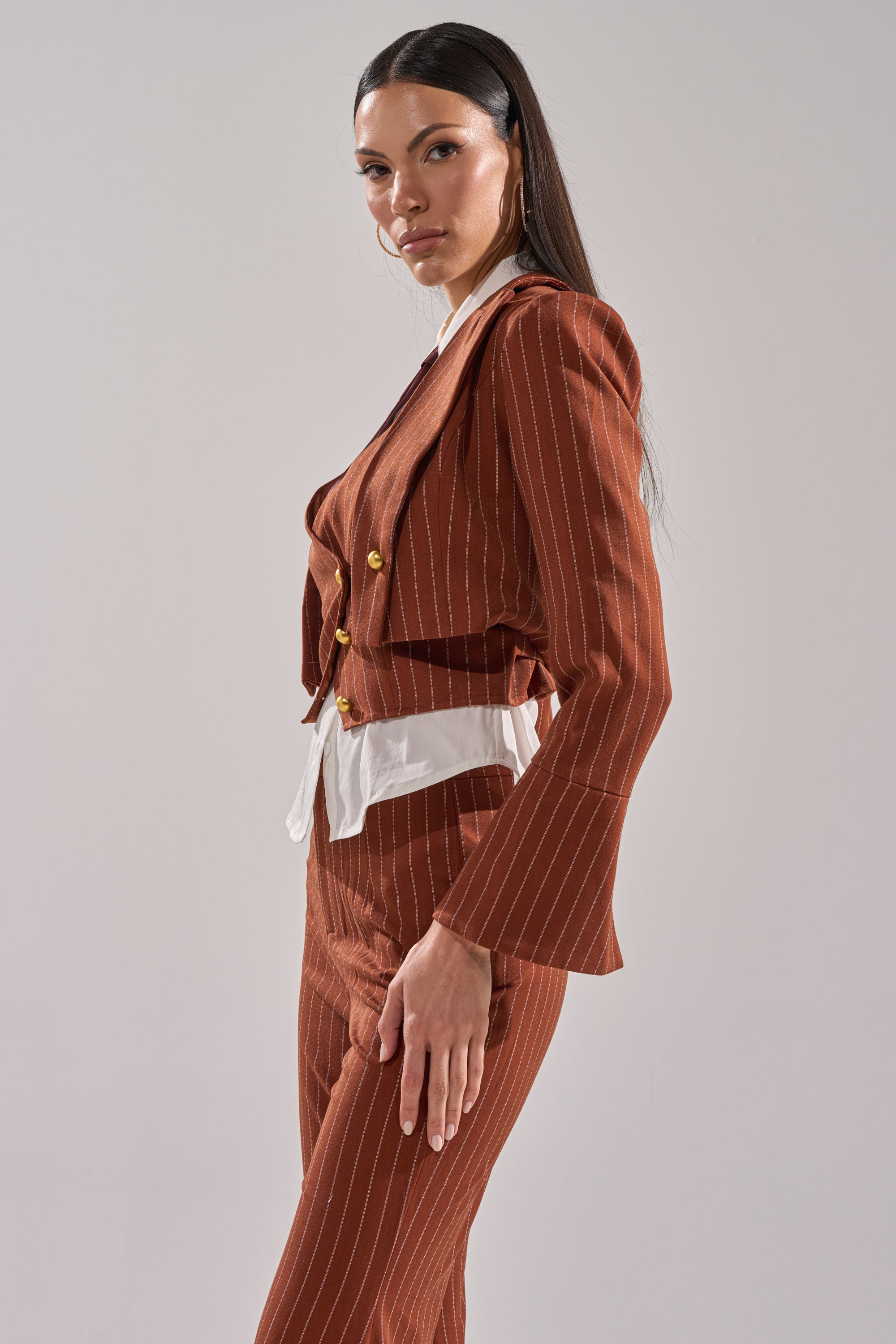 A woman in profile models the BUSINESS FIRST STRIPE BLAZER, a rust pinstripe jacket with gold buttons over a white shirt. She has long straight dark hair and a neutral expression, set against a plain light gray background.