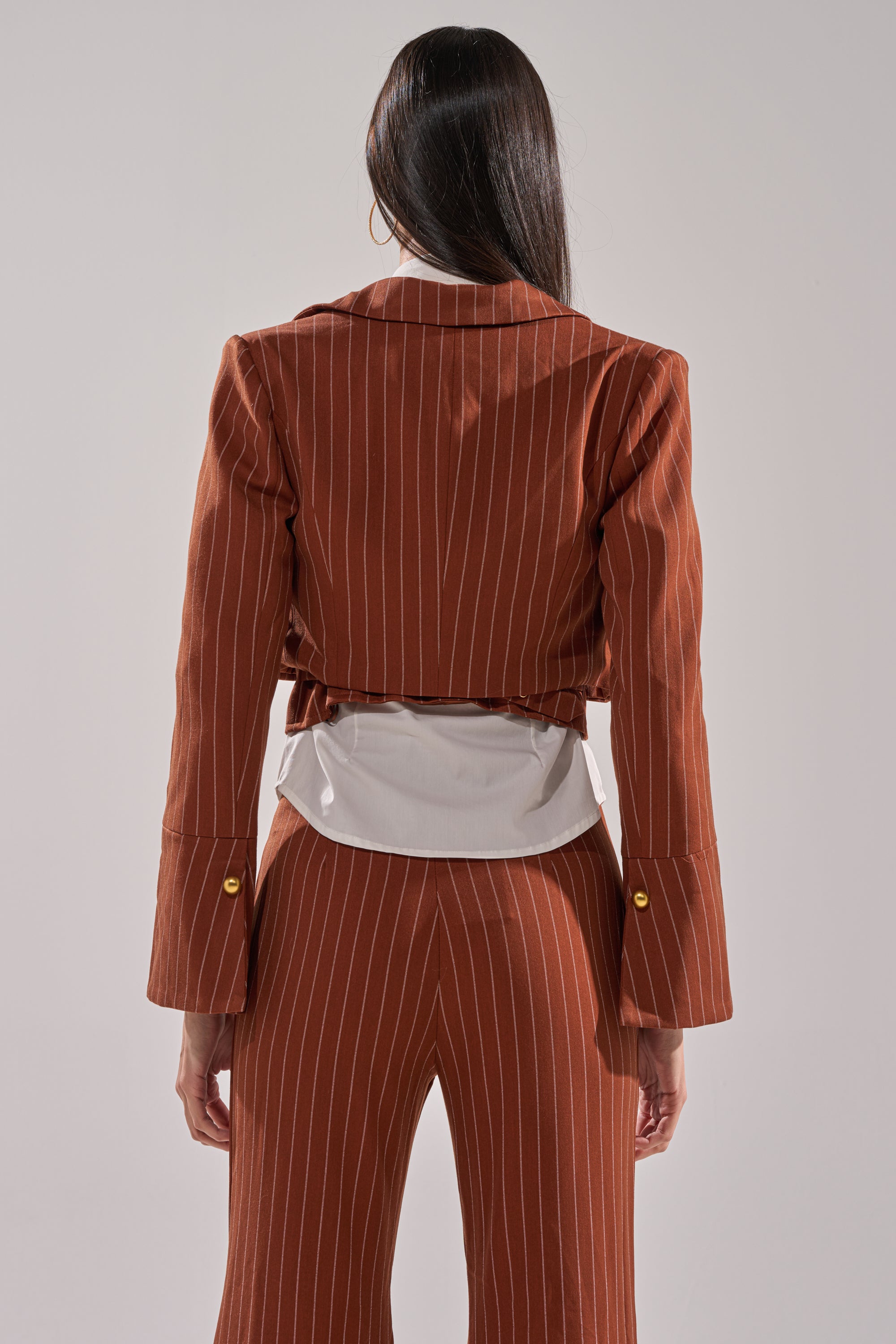 A person with long dark hair stands against a light gray background, wearing the BUSINESS FIRST STRIPE BLAZER over a white shirt, paired with flared pants and a cropped jacket.