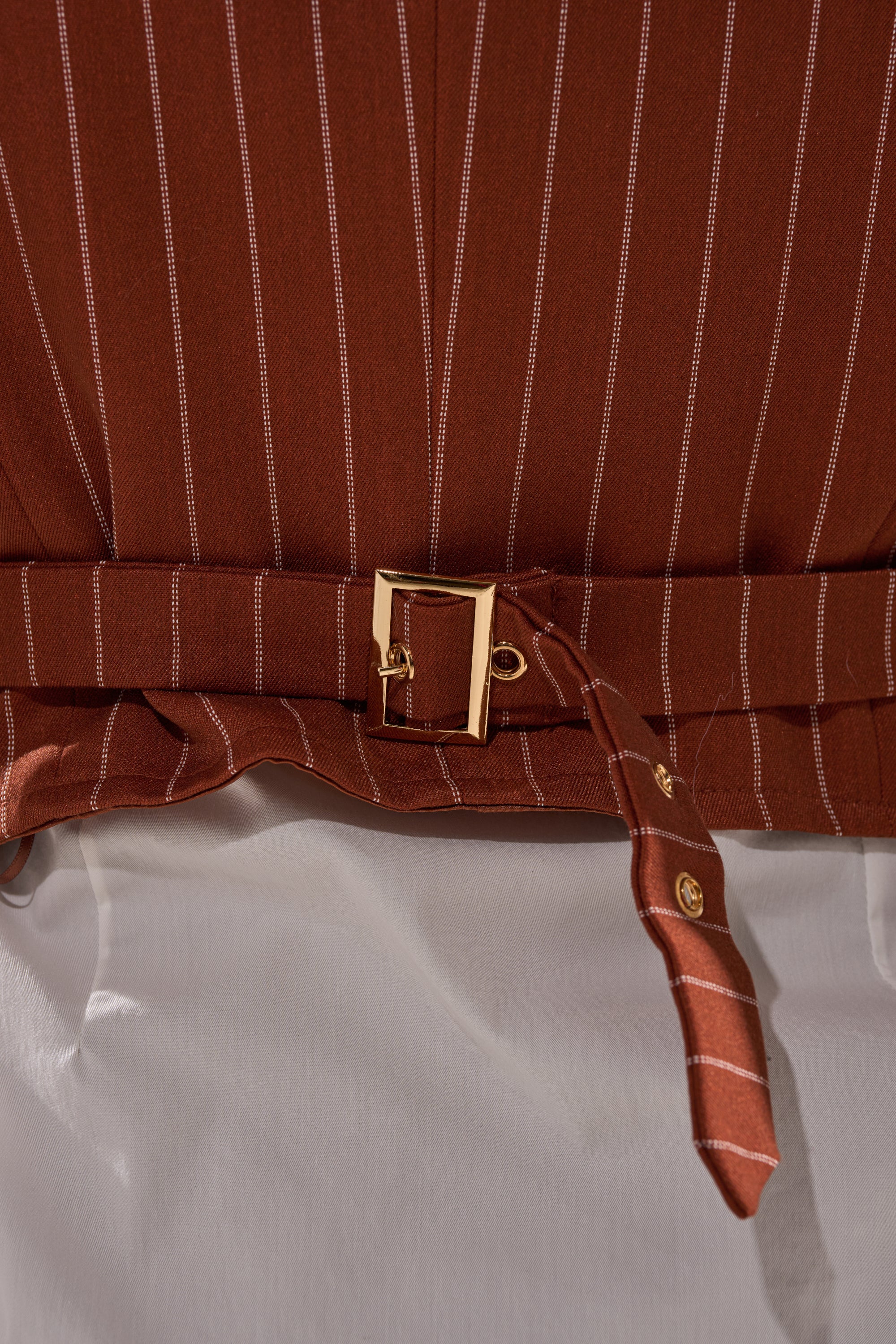 Close-up of the BUSINESS FIRST VEST, a brown pinstripe piece featuring a gold buckle belt at the waist, worn over a white top. Tailored design suggests it's part of a suit ensemble.