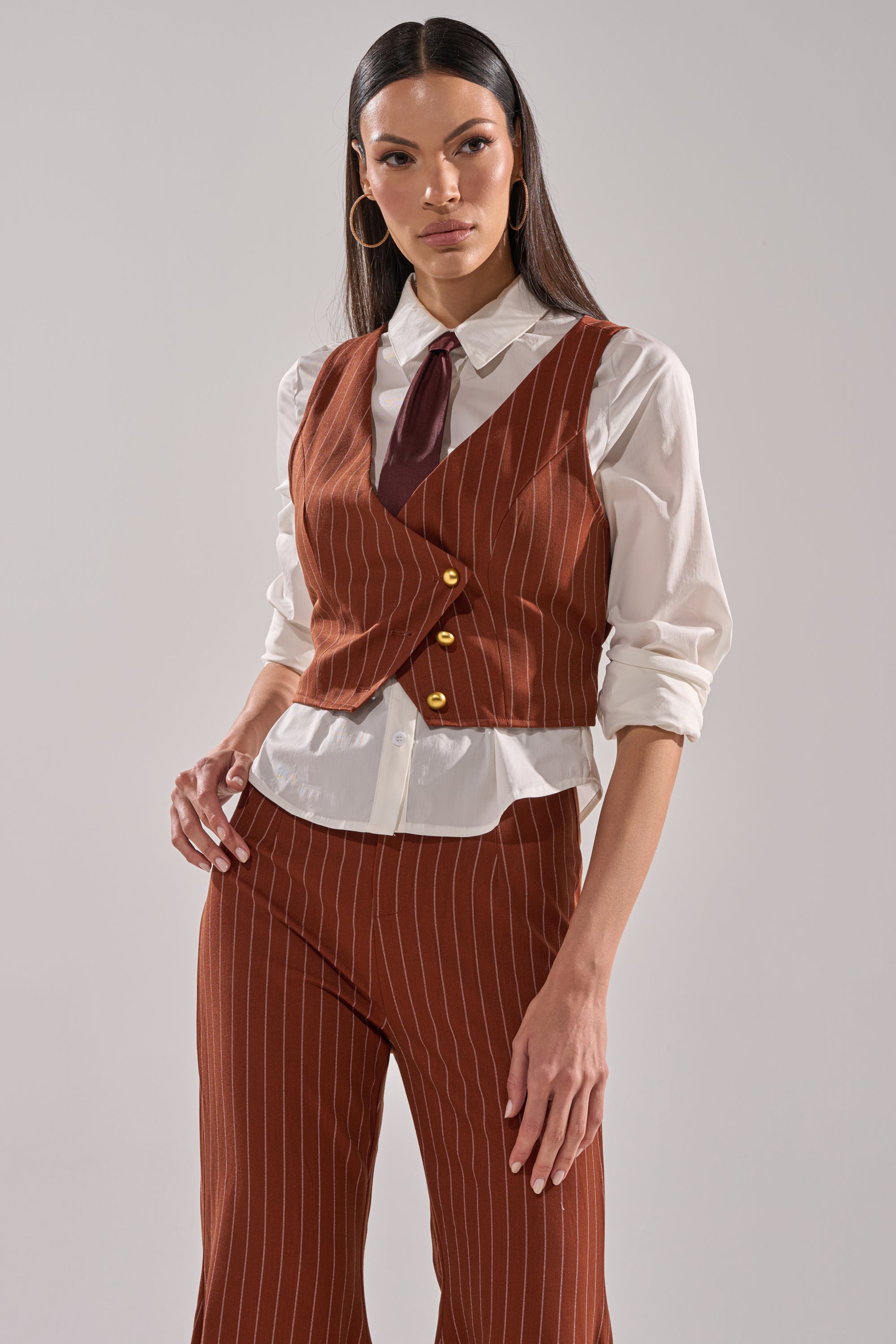 A woman poses confidently in the BUSINESS FIRST VEST with matching pants over a white button-down shirt and dark red tie. She has long straight hair, large hoop earrings, and showcases the vest’s modern cropped fit.